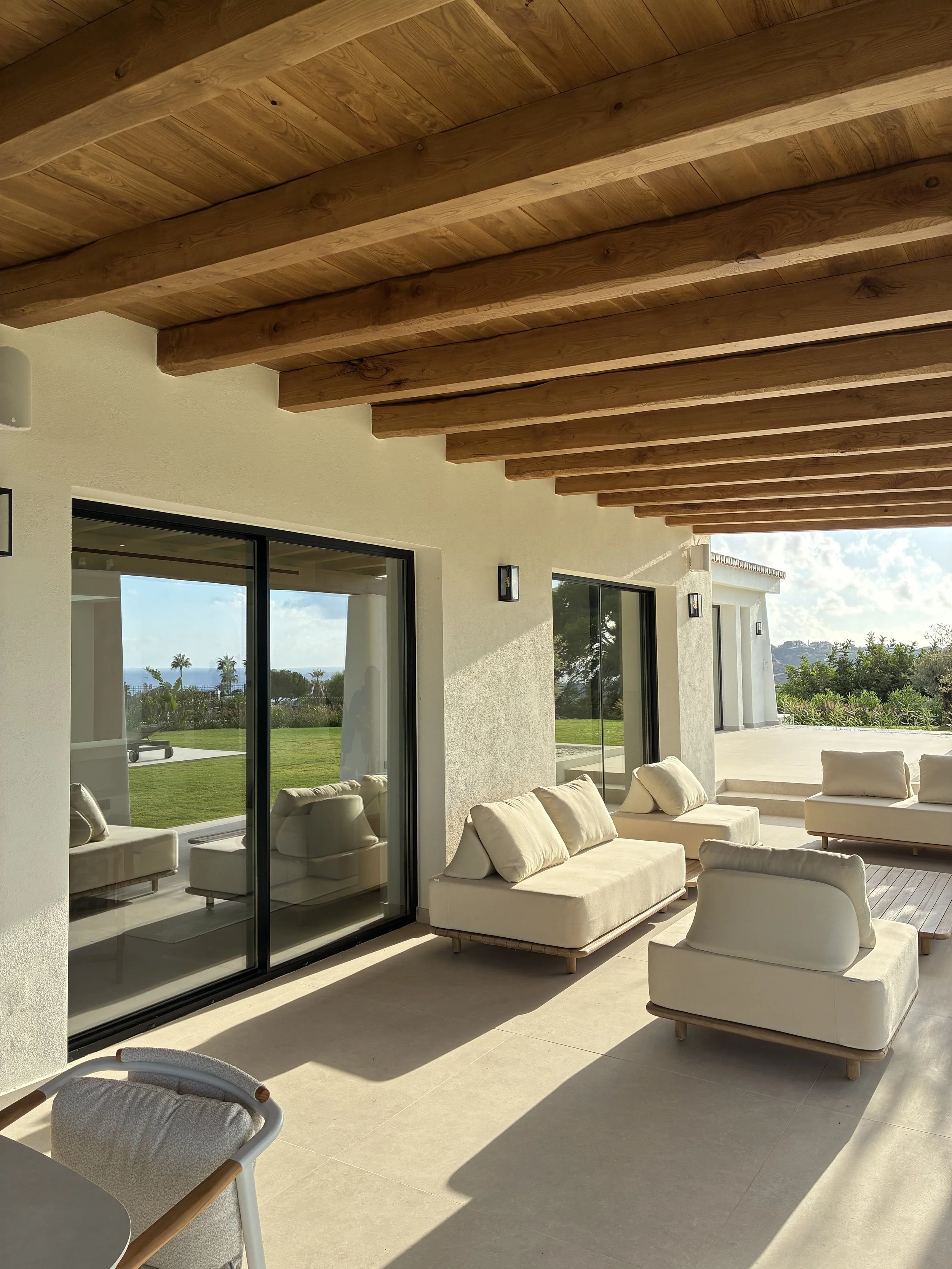 Terraza exterior con sillones beige, paredes blancas, techo de madera, vista al jardín y al paisaje.