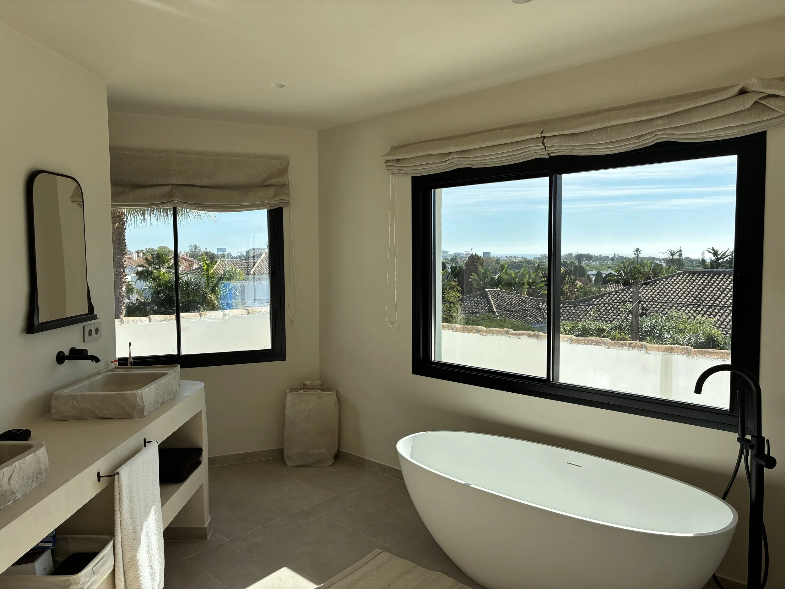 Baño con tina freestanding, dos lavabos de piedra, ventanas grandes con vista a casas, árboles y cielo, decoración minimalista en tonos neutros.