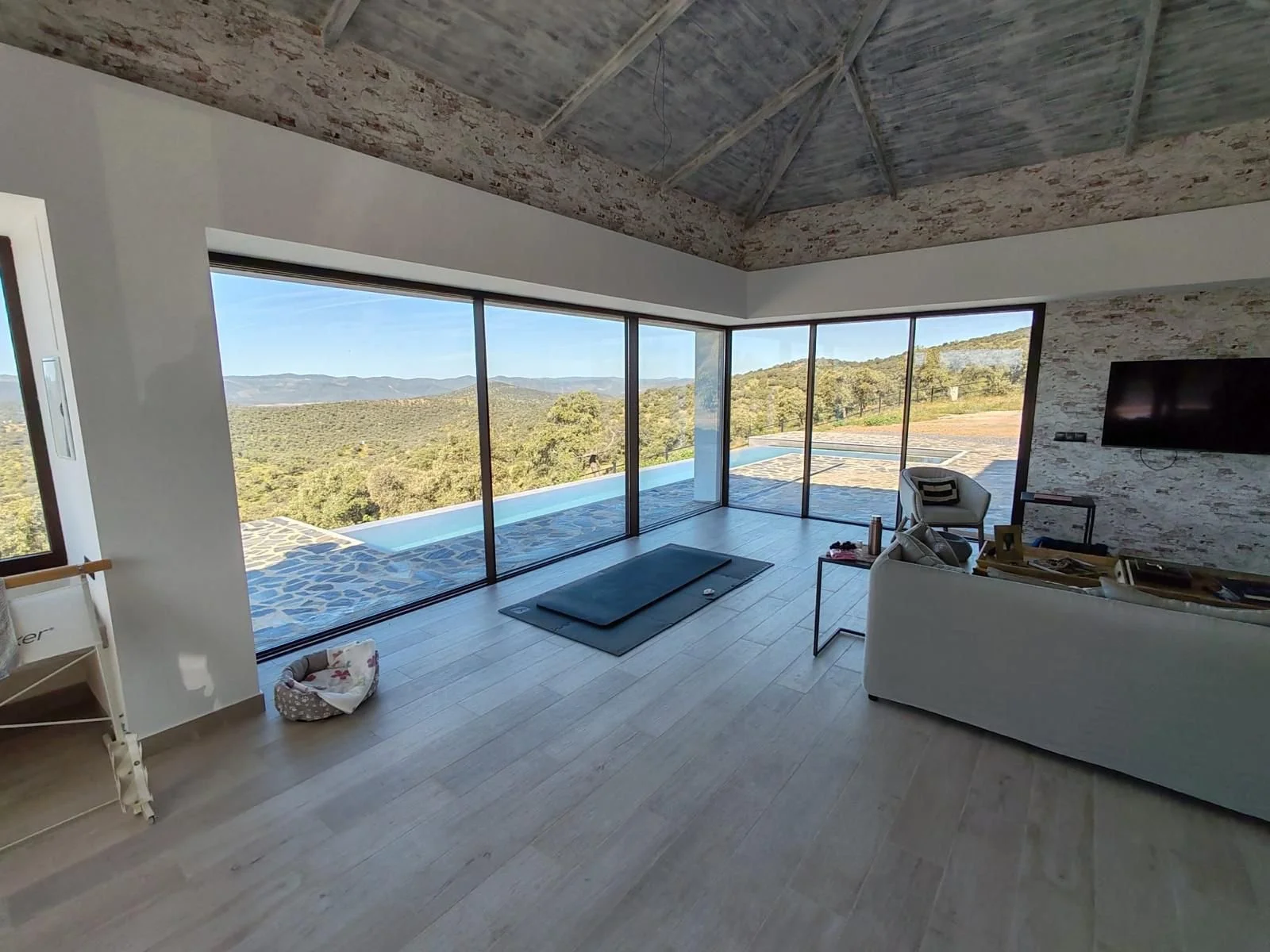 Vista de una sala moderna con ventanas de vidrio grandes, mostrando un paisaje de montañas y árboles. Hay un sofá, una silla, una mesa de centro y un televisor en la pared.