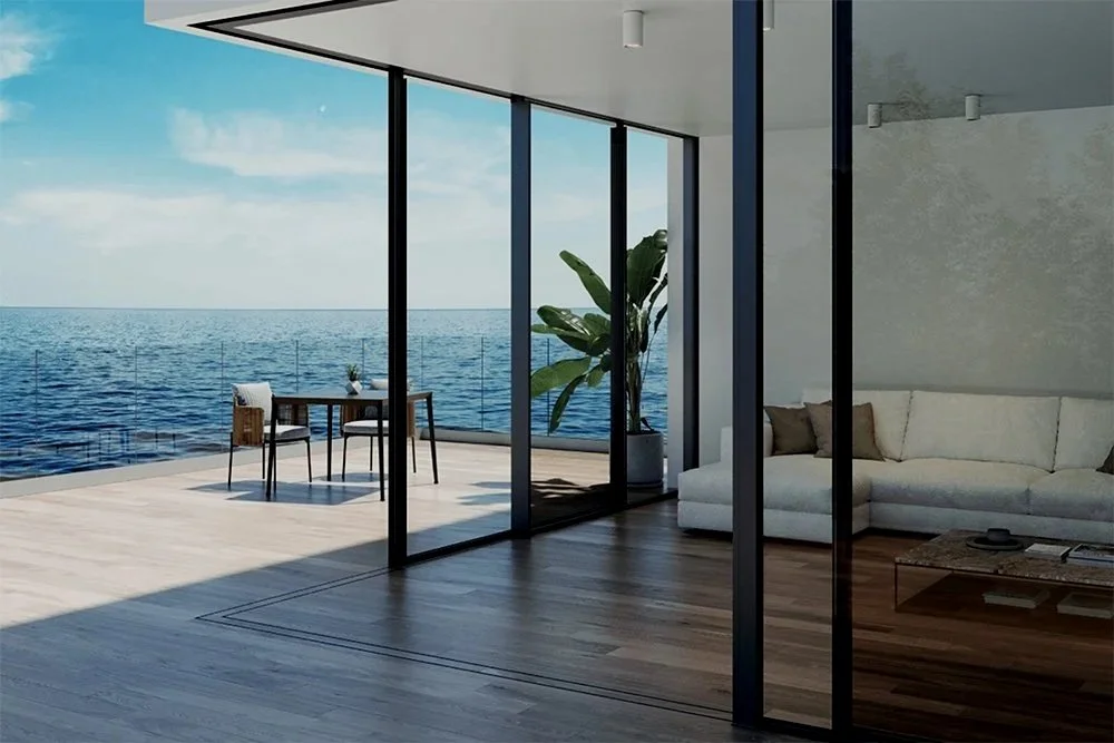 Vista moderna de una sala con paredes de cristal que da a una terraza con vista al mar, con sofá, mesa y sillas en un ambiente luminoso y abierto.