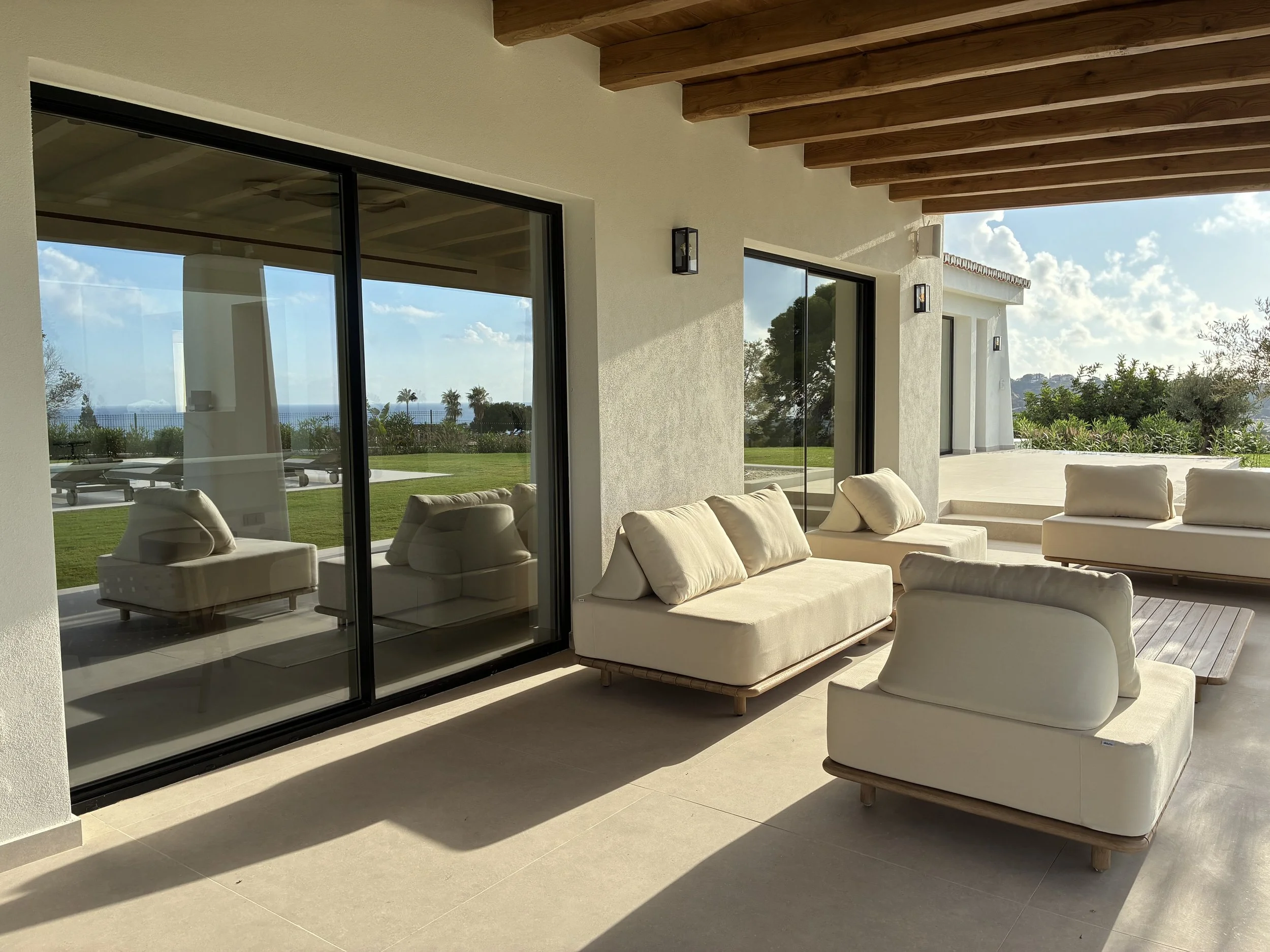 Sala de estar exterior con sillones blancos y grandes ventanales, vista a un jardín y al mar, en un día soleado.