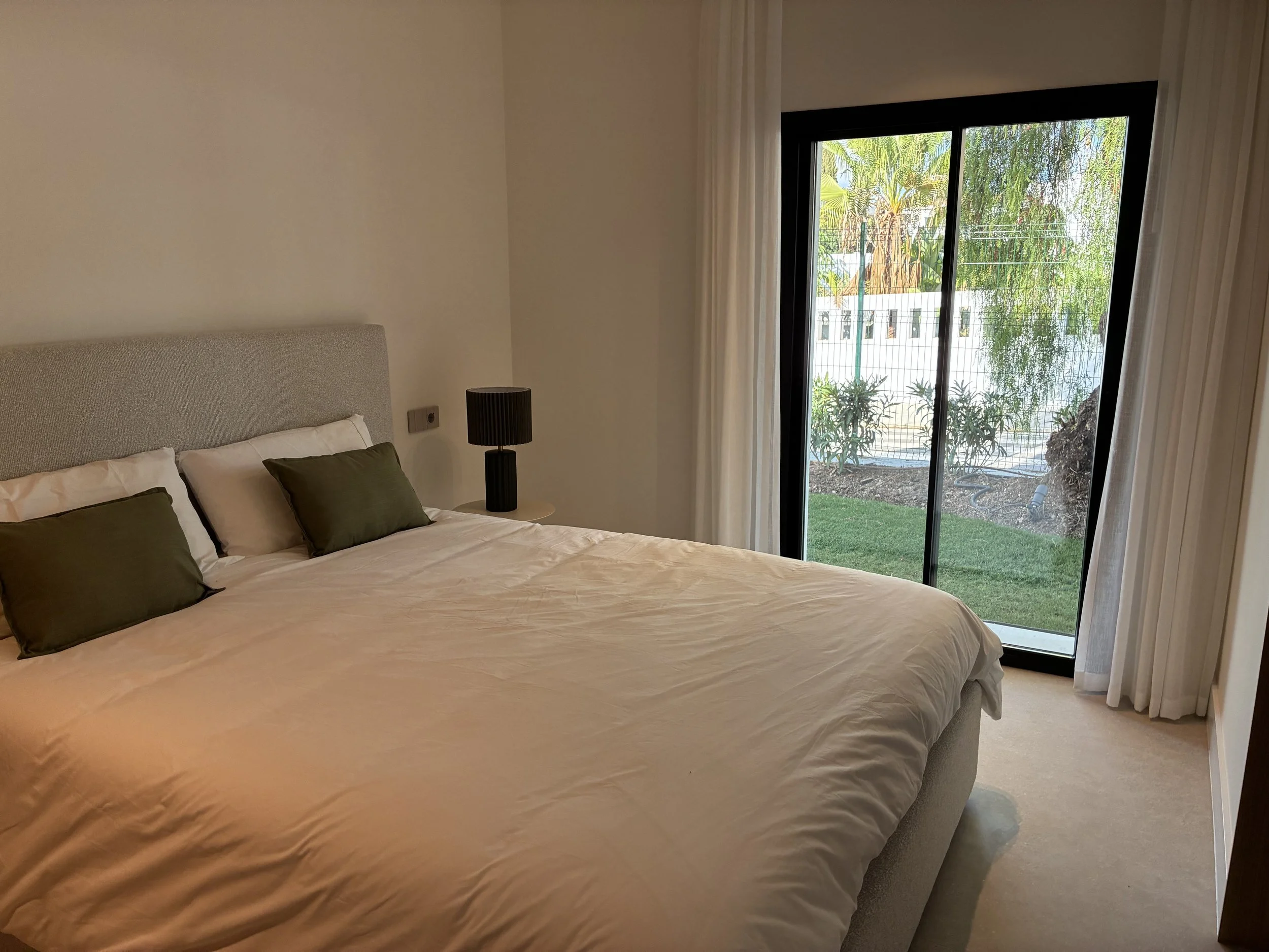 Habitación con cama grande, almohadas blancas y verdes, lámpara negra y vista a un jardín con césped y plantas, vista a través de puertas corredizas de vidrio con cortinas blancas.