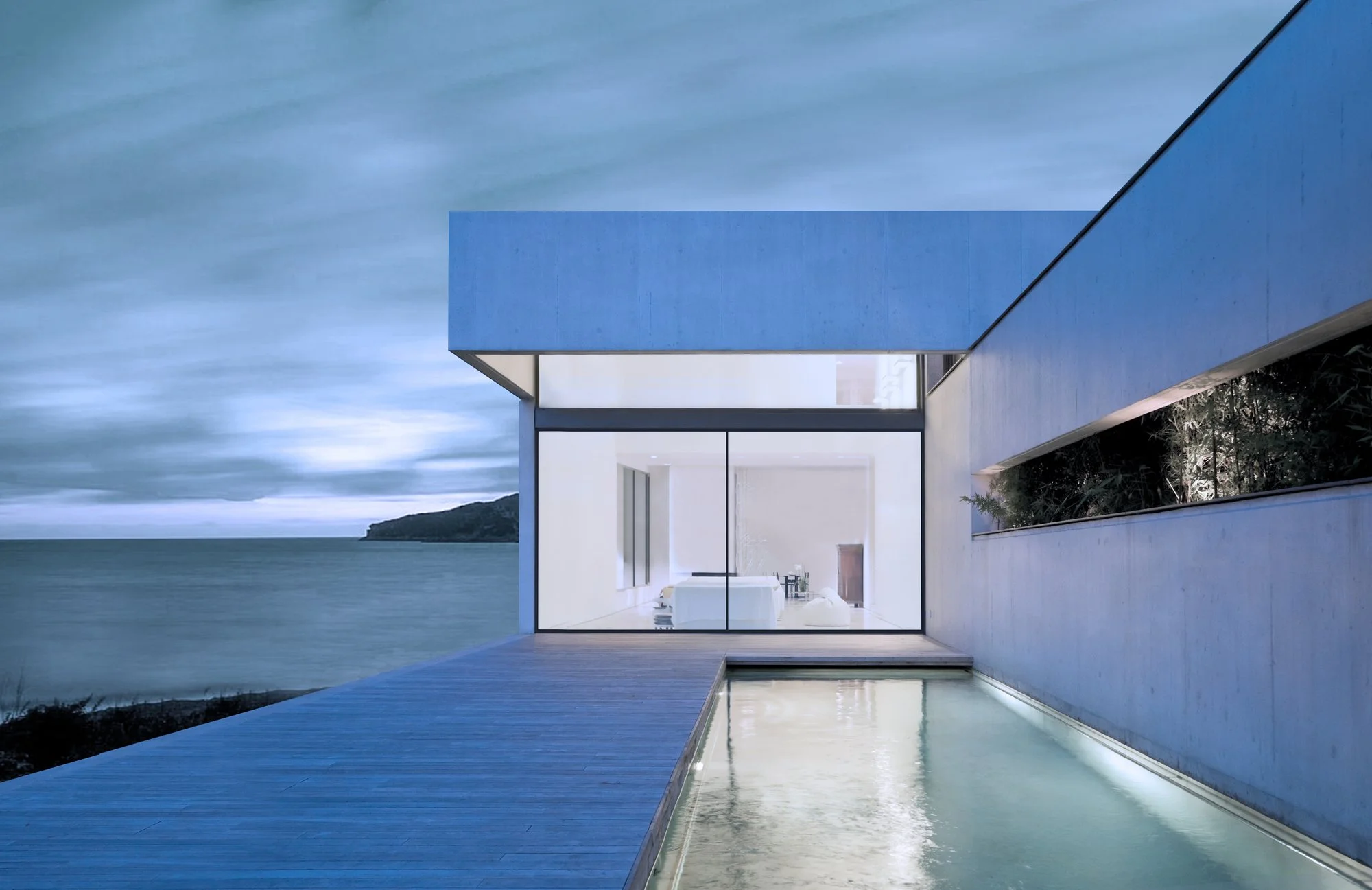 Modern house with large glass windows and a pool overlooking the ocean under a cloudy sky.