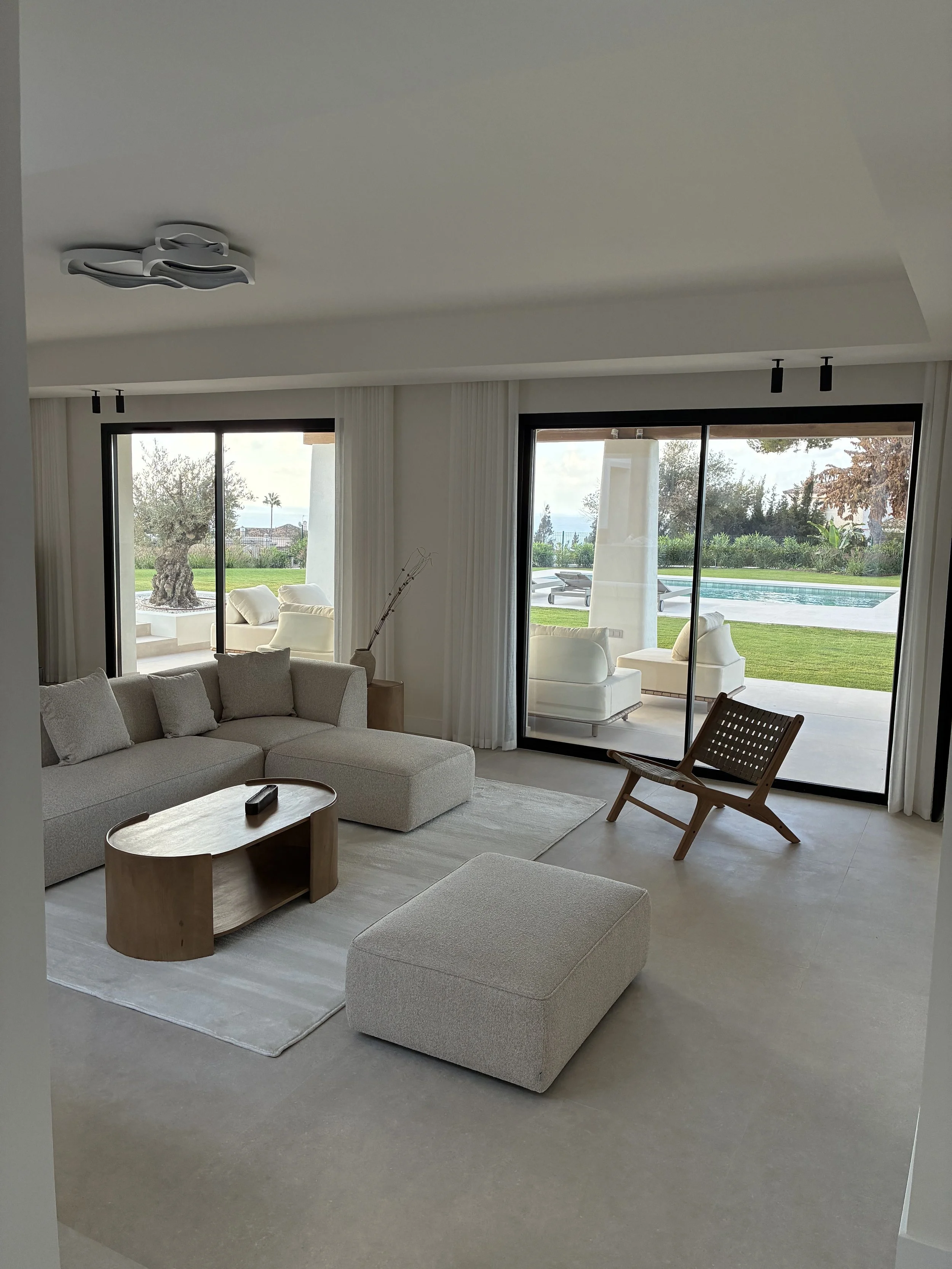 Sala moderna con sillón beige, otomana, mesa de madera, silla de mimbre, y vista al jardín y piscina al exterior.