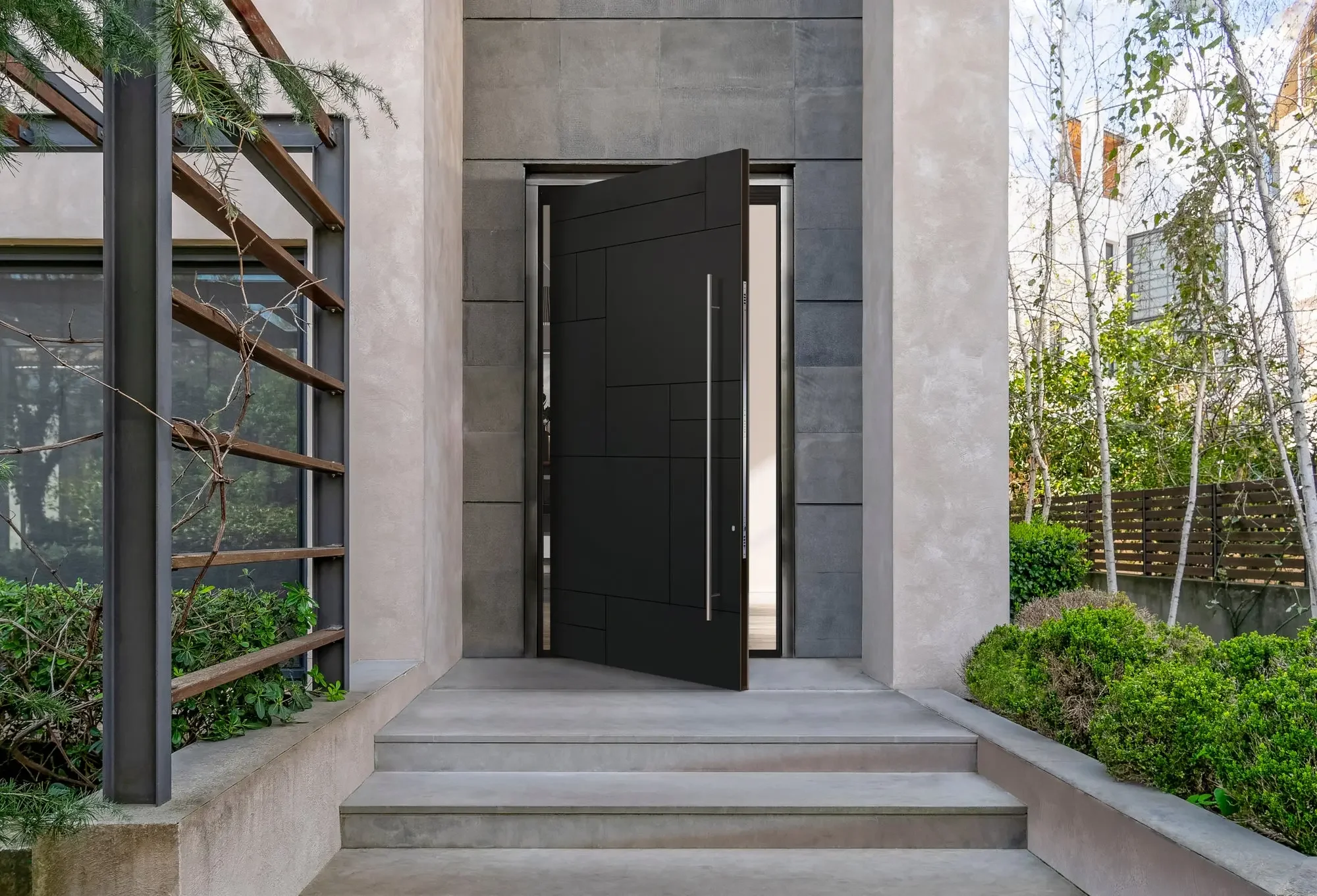 Entrada moderna a una casa con puerta negra y escalones de concreto, rodeada de plantas y enrejado de madera en el jardín.