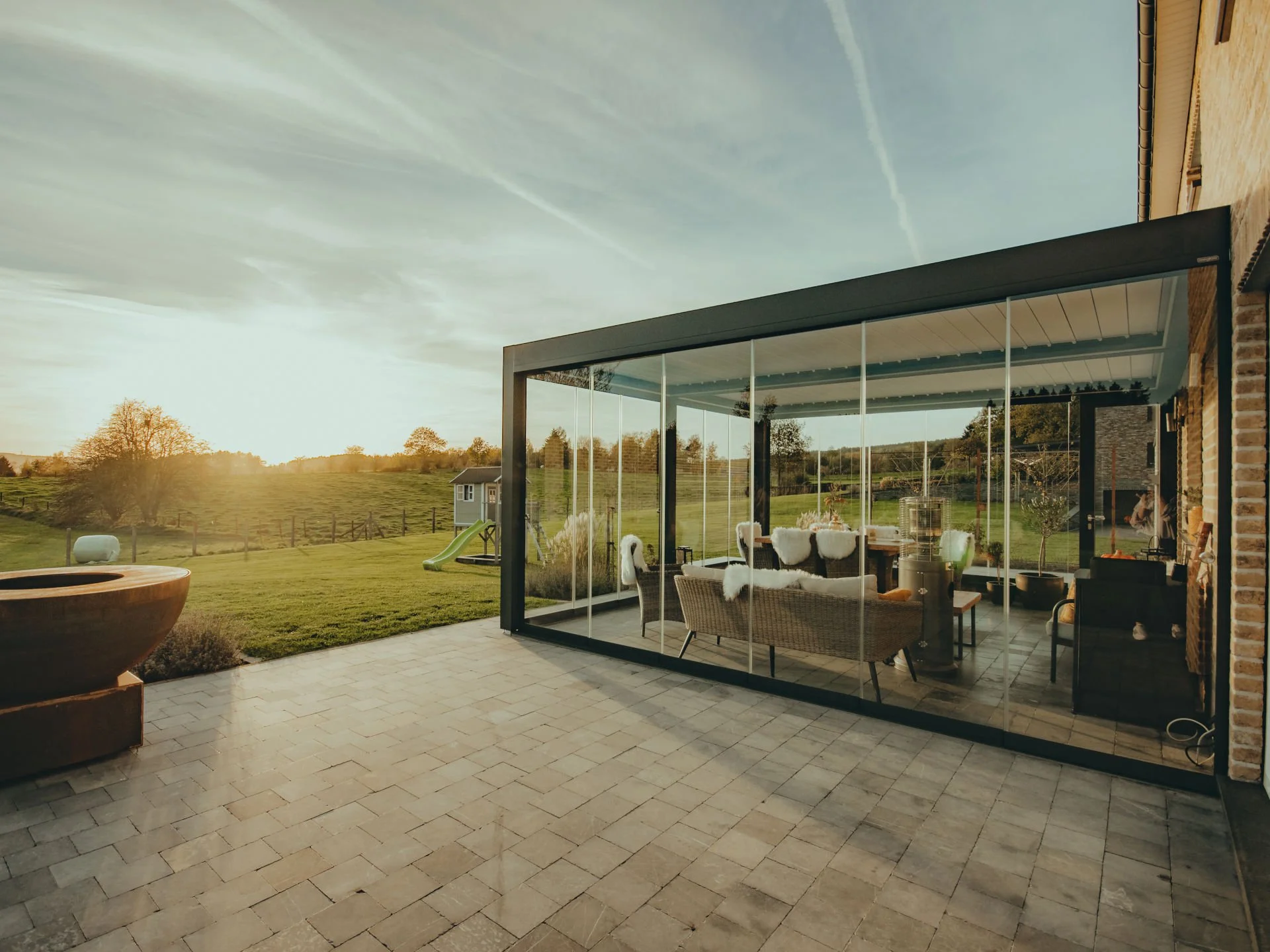 A glass-enclosed patio attached to a house, with outdoor chairs, a small table, and a heater inside. The patio overlooks a grassy yard with trees, a slide, and a small shed, under a partly cloudy sky at sunset.