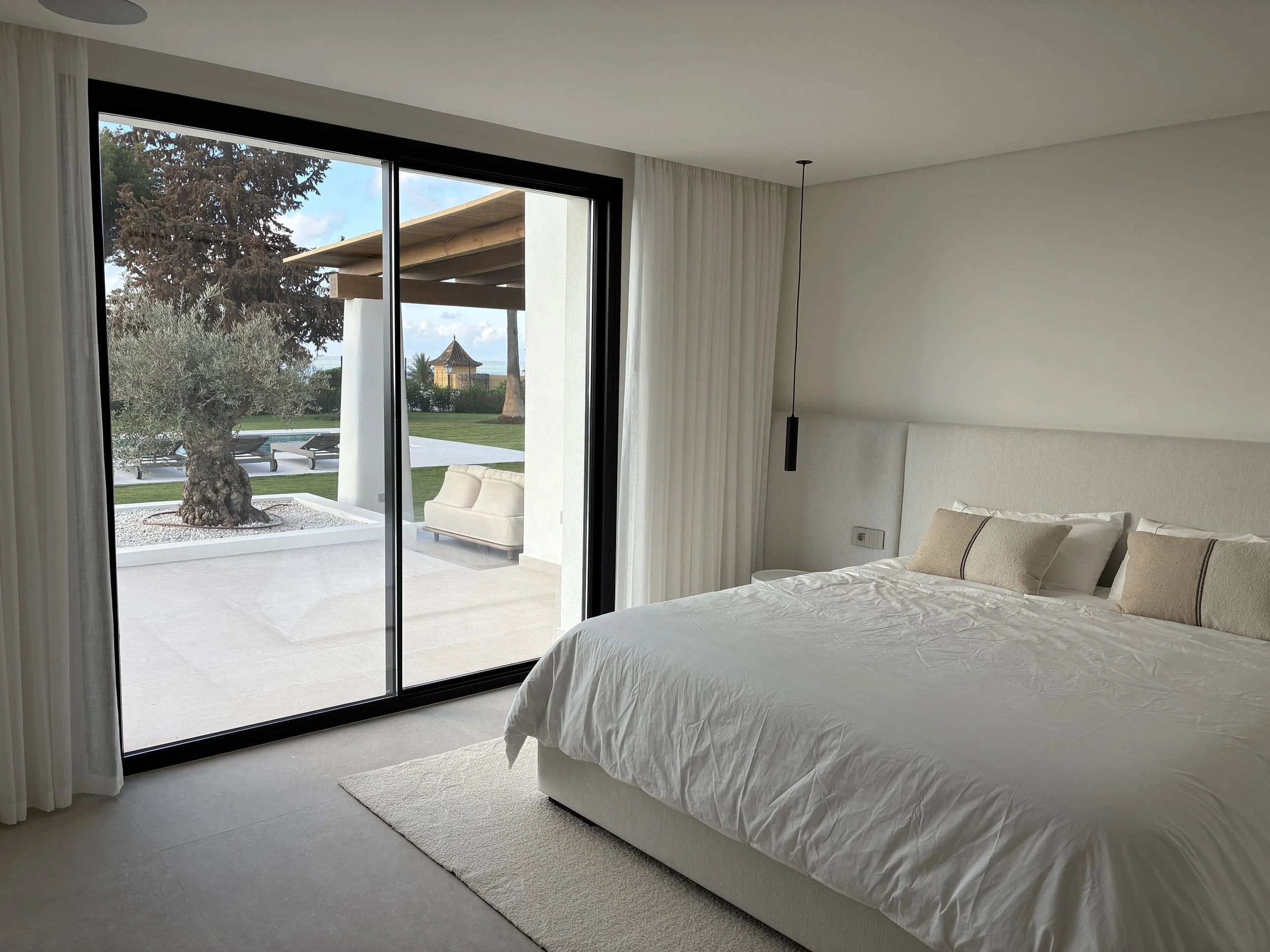 Habitación moderna con cama blanca, vista a un patio exterior con árbol, césped y tramos de piedra, a través de una gran puerta de cristal corrediza.