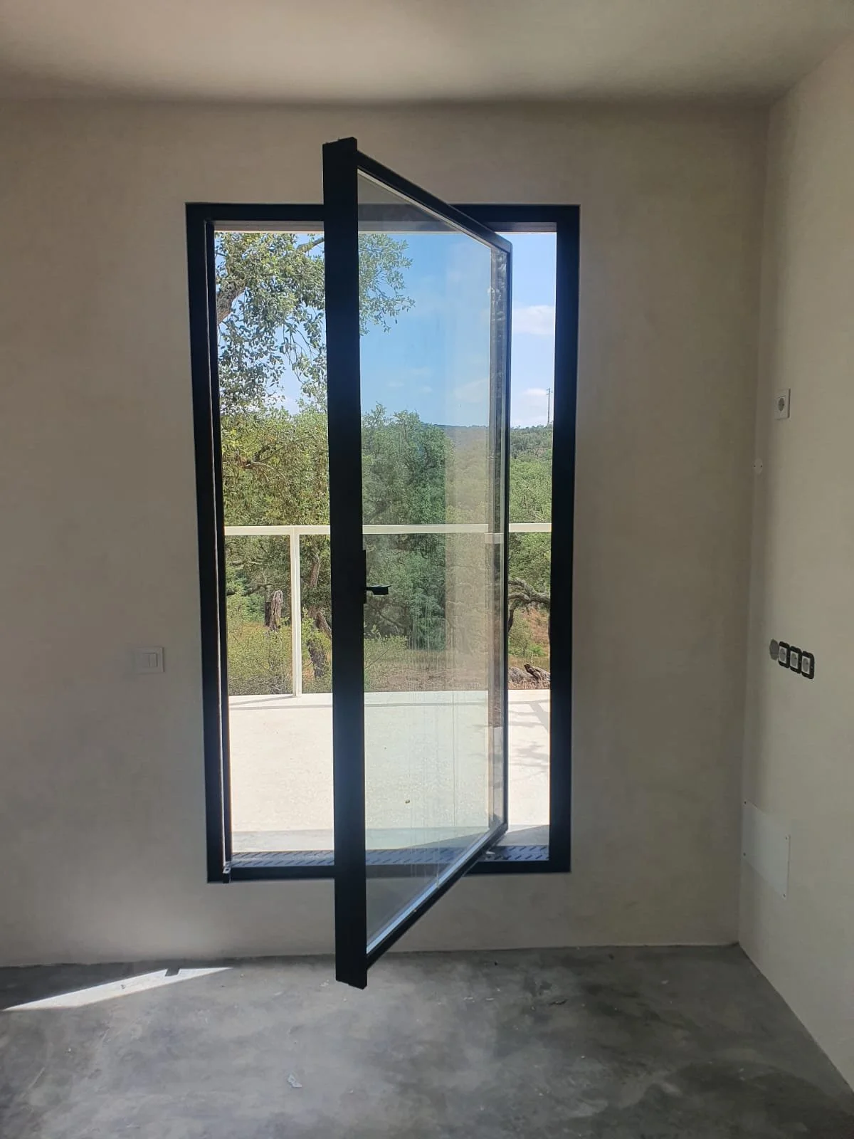 Ventana de vidrio con marco negro en una pared de una habitación vacía, con vista a un área exterior con árboles y cielo azul.