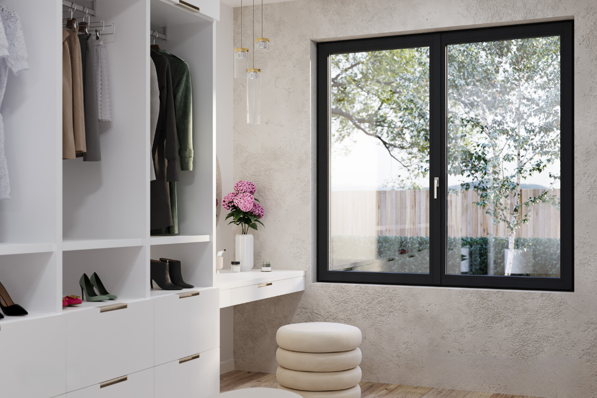 A walk-in closet with open wardrobes displaying clothes and shoes, a white vanity with a pink flower arrangement, a cream cushioned stool, and a large window showing outdoor trees and a wooden fence.