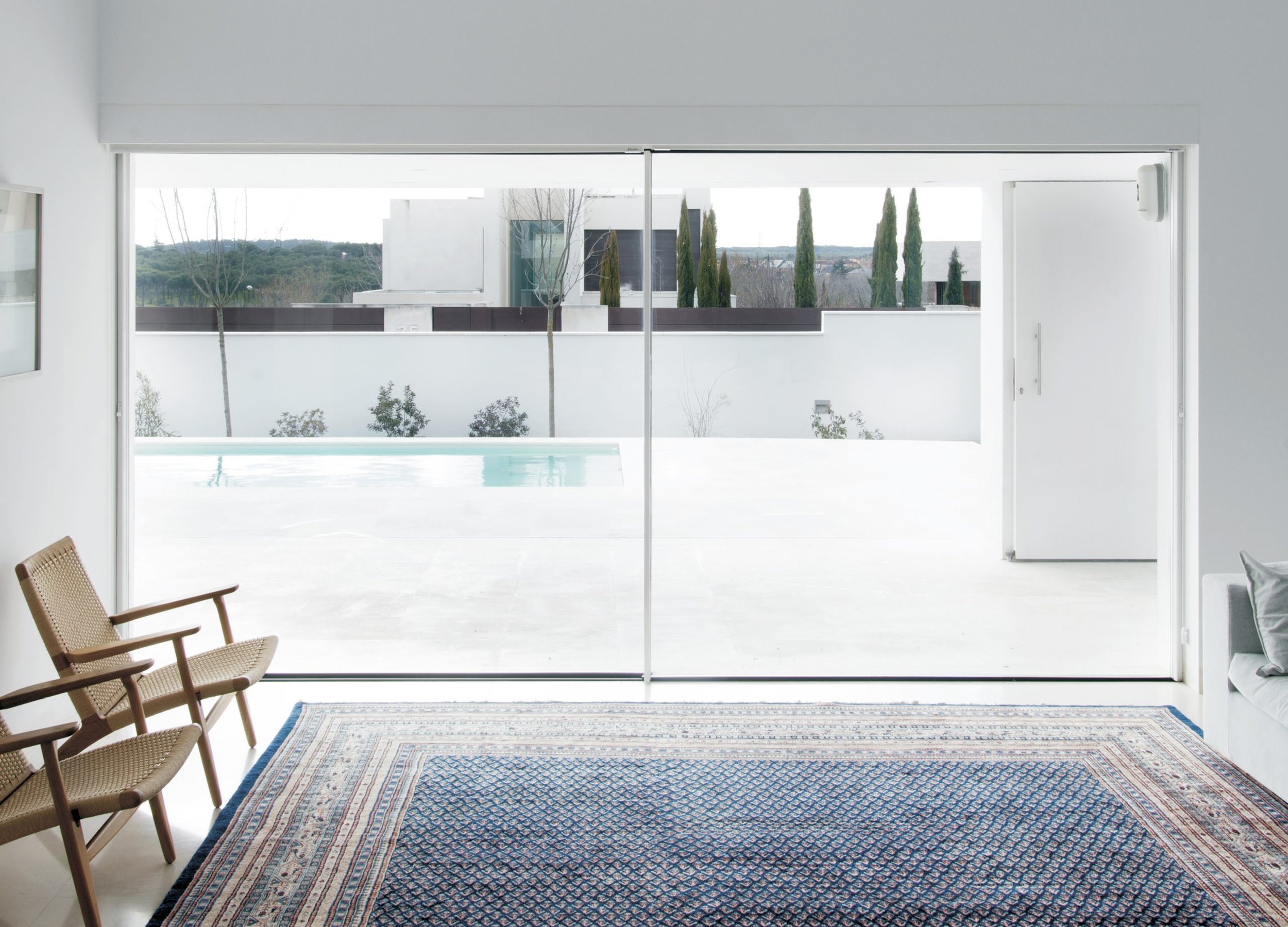 Modern living room with large sliding glass doors opening to a backyard with a pool, white walls, a patterned rug, and minimalist furniture.