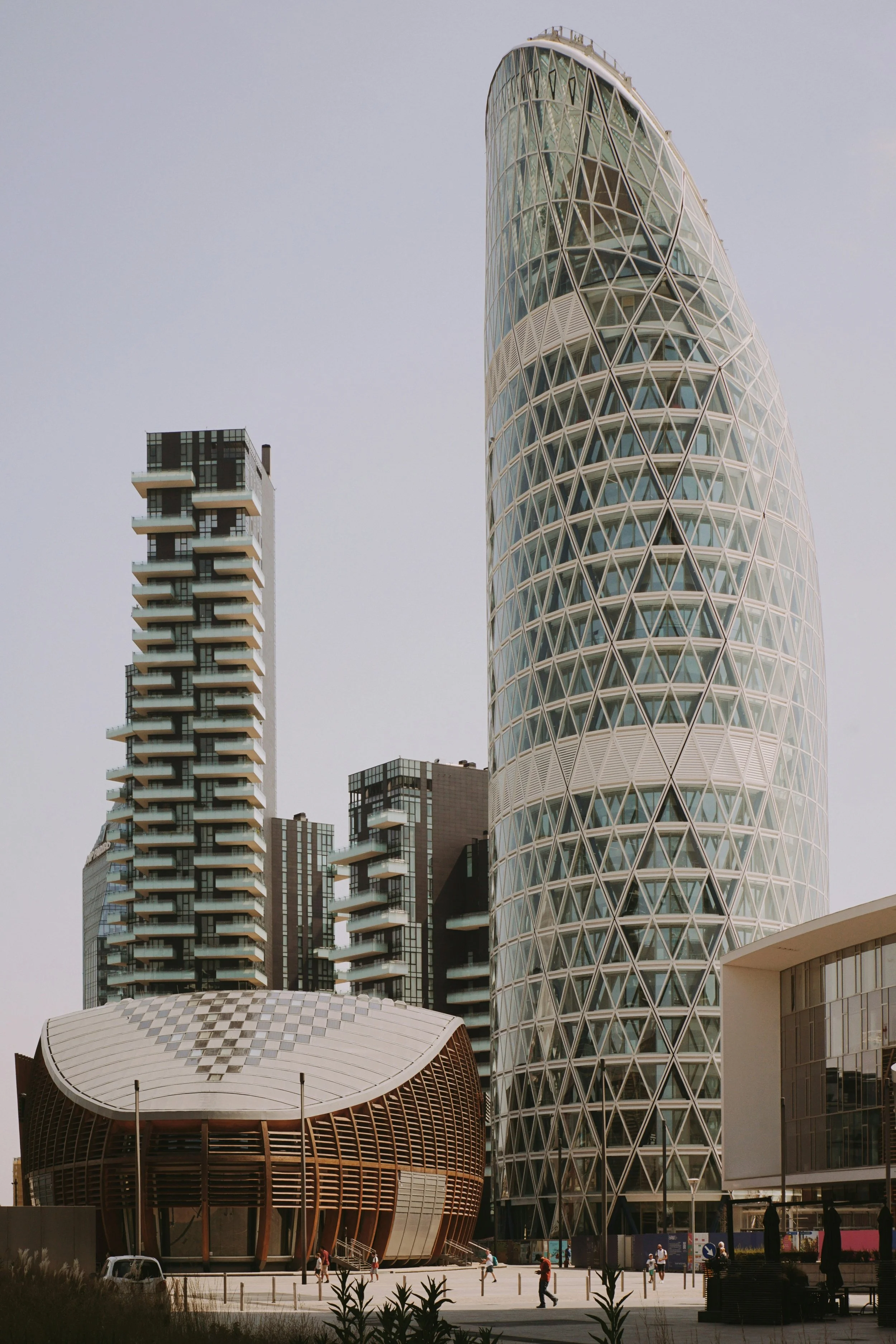 Modern cityscape with unique architectural buildings, including a tall, curved glass skyscraper and a round, wooden building at street level with people walking nearby.