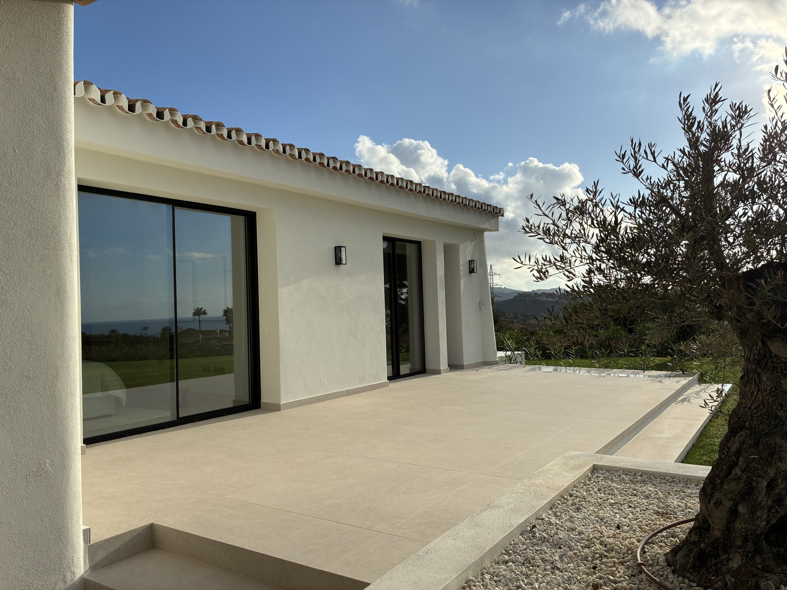 Vista de una casa moderna blanca con ventanas de vidrio y puertas corredizas, un patio de cemento, un árbol y un paisaje montañoso con cielo despejado y algunas nubes.