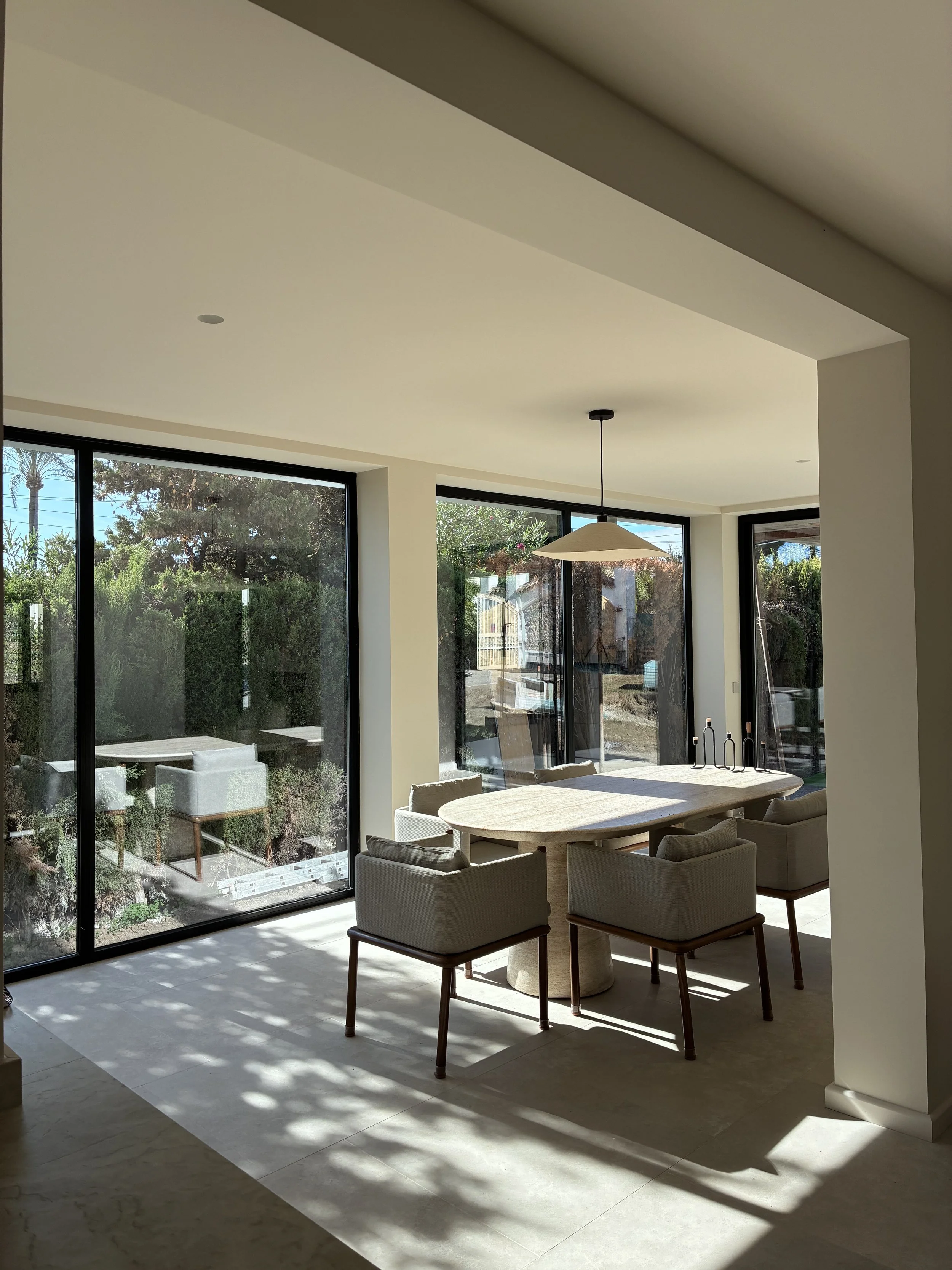 Comedor con mesa de madera y sillas de tela, grandes ventanas de vidrio que dejan entrar luz natural, vista al jardín exterior, lámpara colgante, ambiente moderno y minimalista.