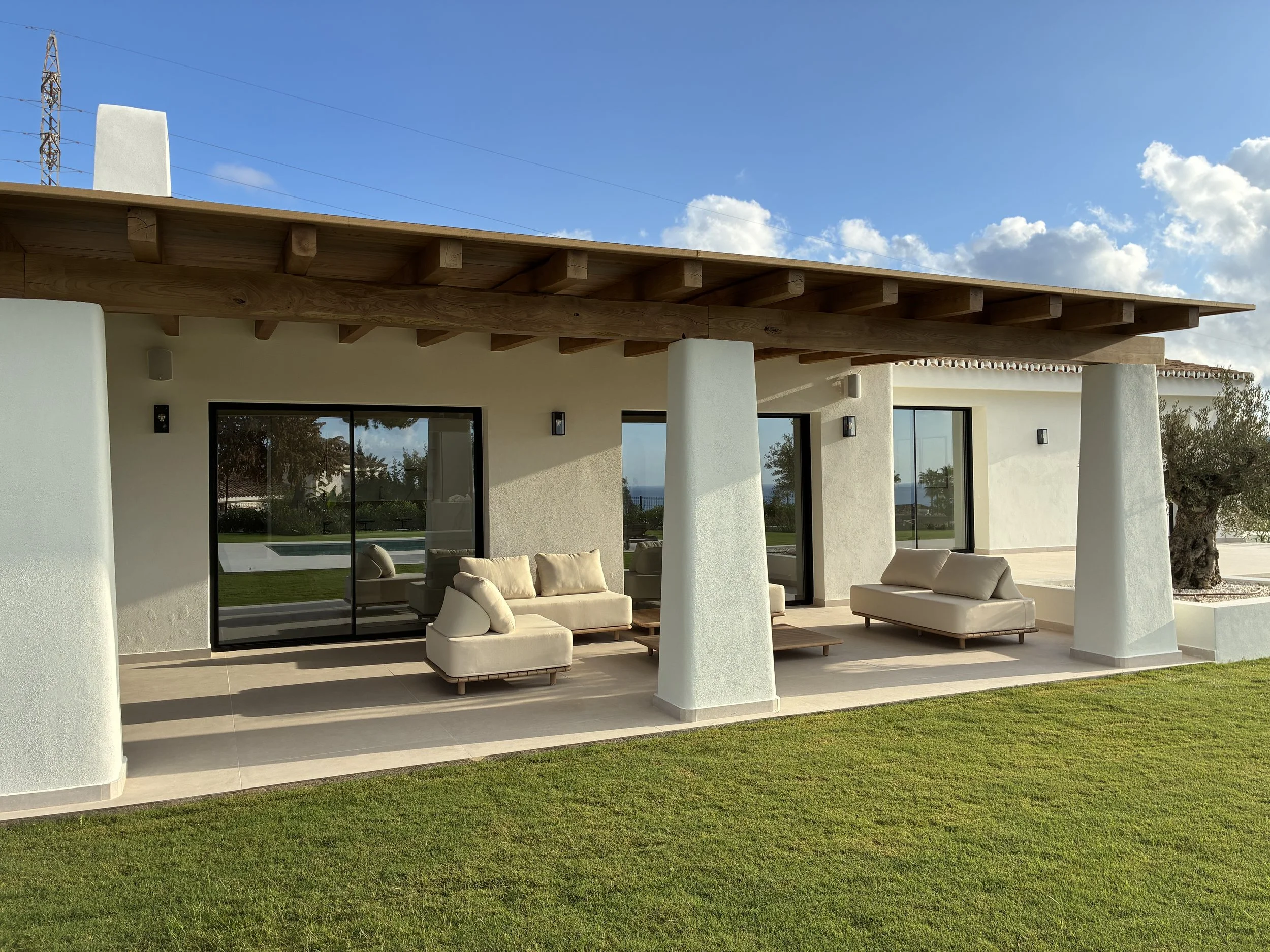 Casa moderna de una planta con patio cubierto, muebles de exterior blancos y vista al jardín, en un día soleado.