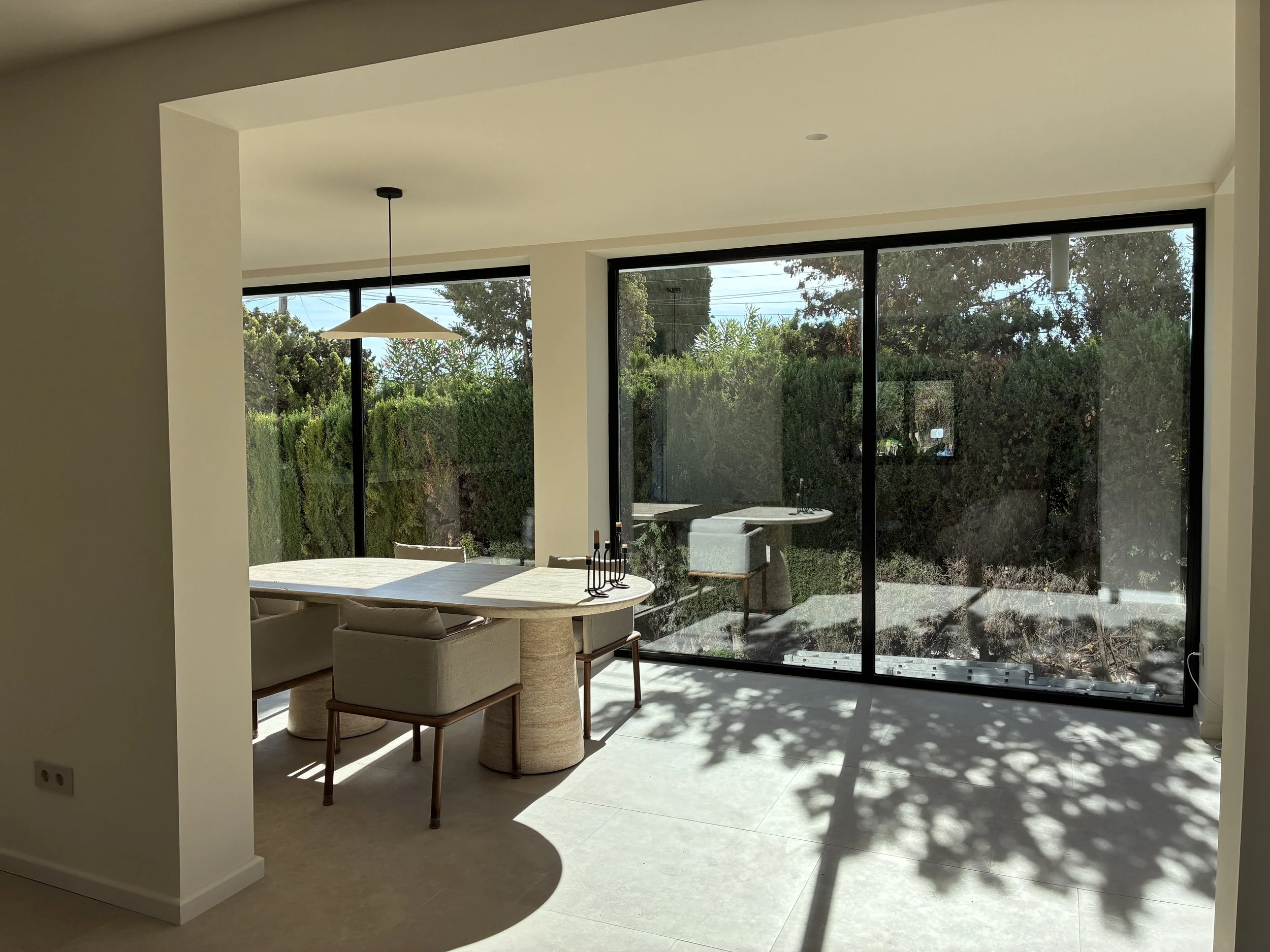Sala de comedor con mesa, sillas y grandes ventanas que dejan entrar luz natural y muestran un área exterior con árboles.
