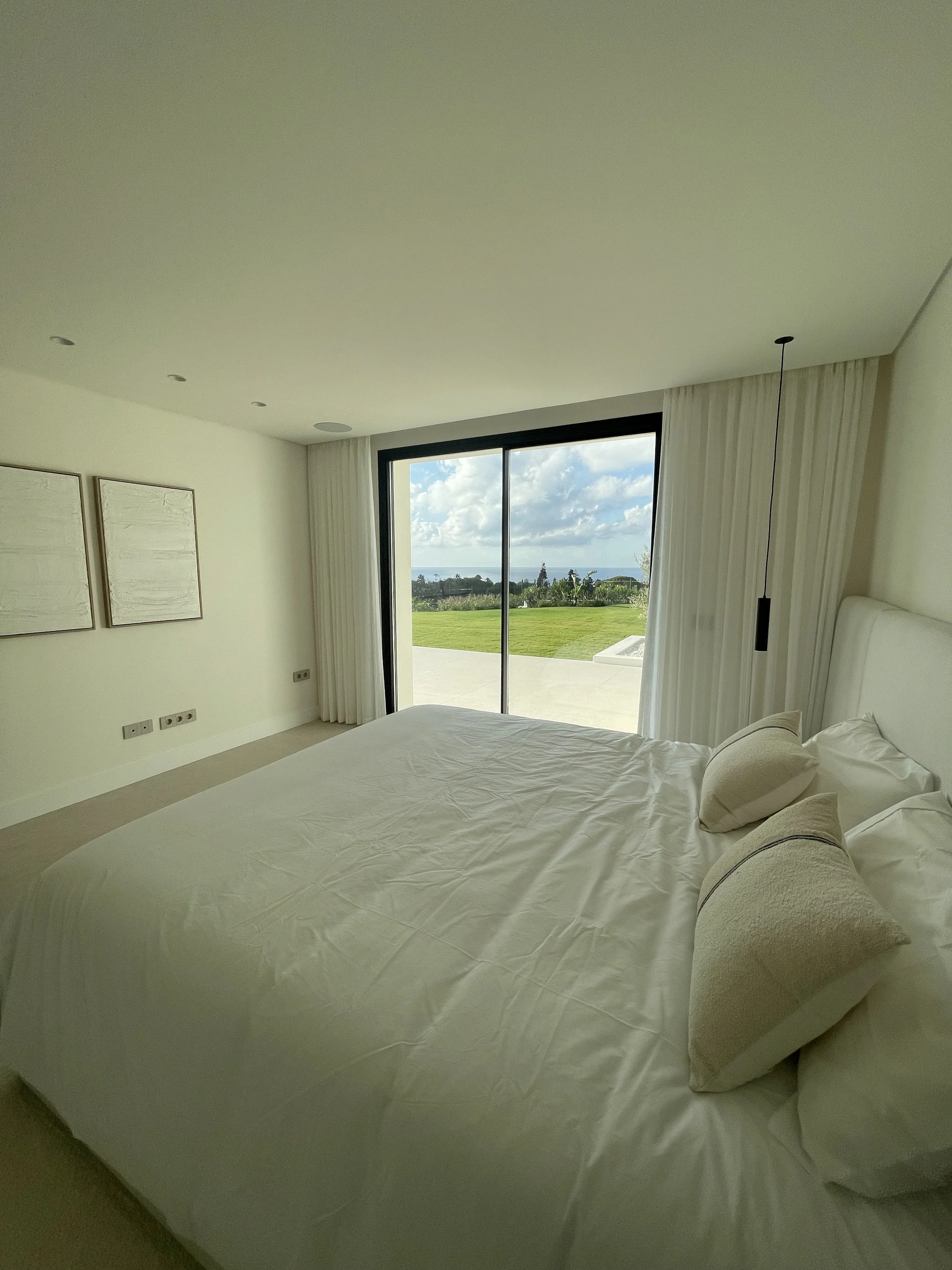 Habitación moderna con cama grande, sábanas blancas y cojines, vista al jardín y al mar a través de un gran ventanal con cortinas blancas.