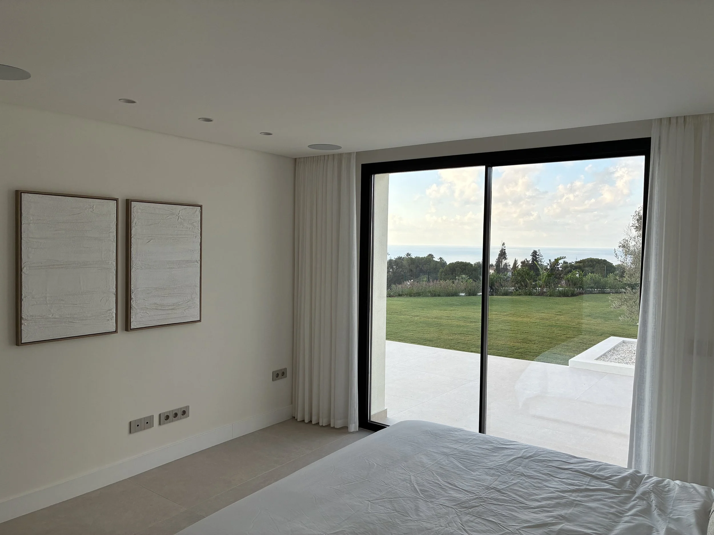 Habitación moderna con cama, ventanas grandes con vista al jardín y al mar, decorada en tonos blancos y neutros.