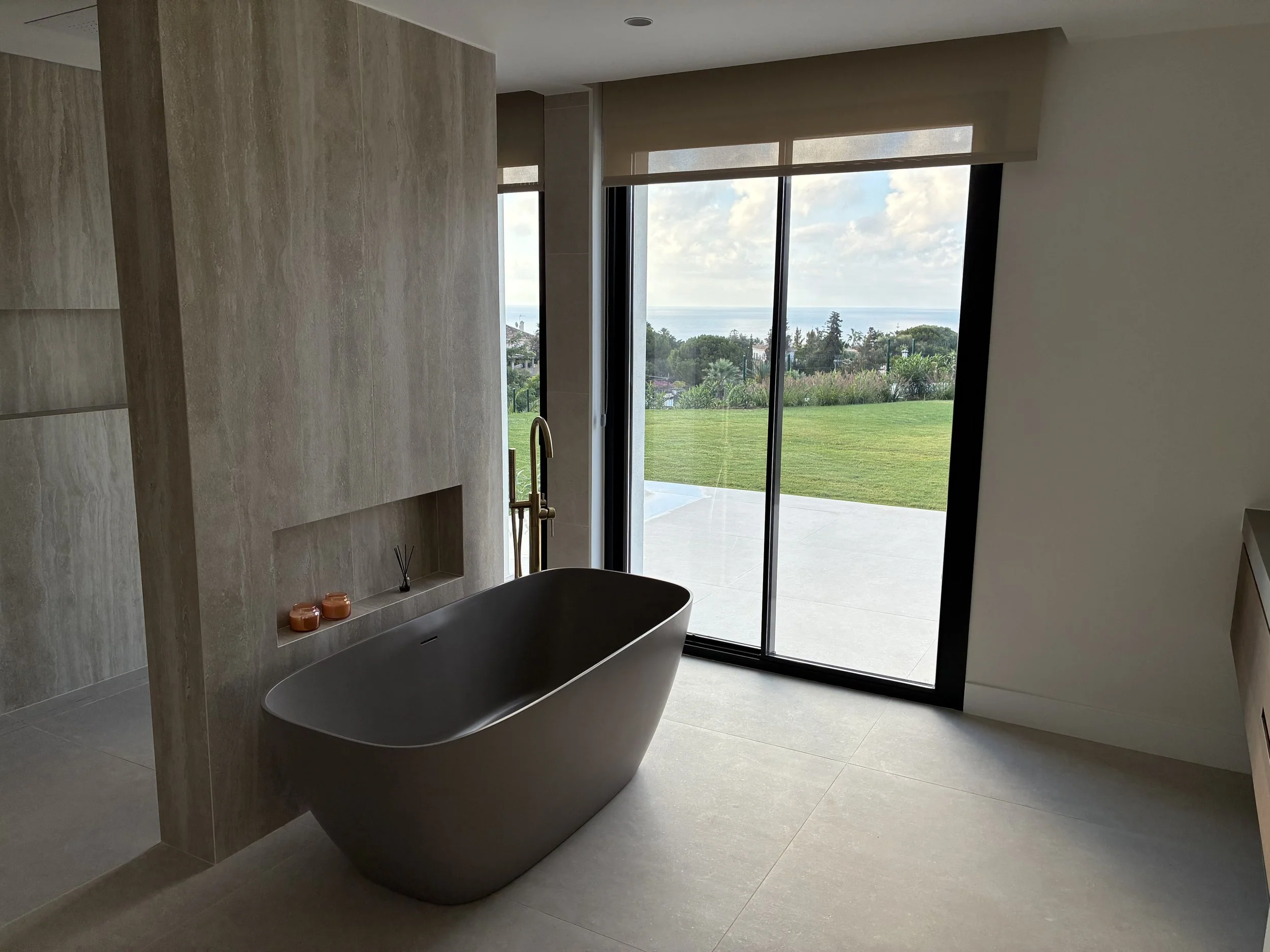 Baño moderno con bañera de pie, vista a un jardín exterior con césped y árboles a través de ventanas grandes.