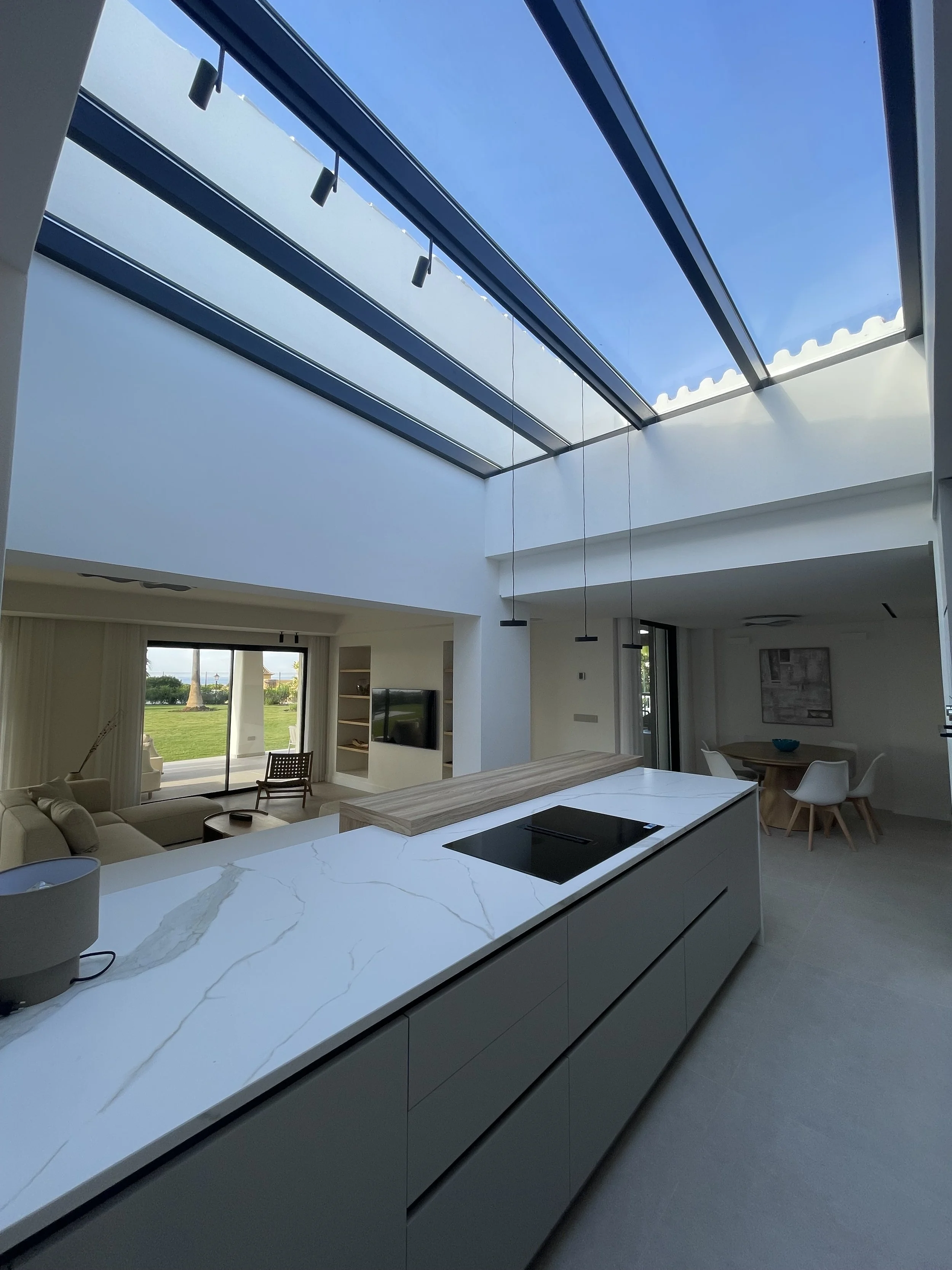 Interior de una cocina moderna con una gran ventana de techo que permite la entrada de luz natural, mostrando una vista del cielo azul. La cocina tiene una isla con encimeras blancas de mármol y una estufa eléctrica, y en el fondo se observa un área 