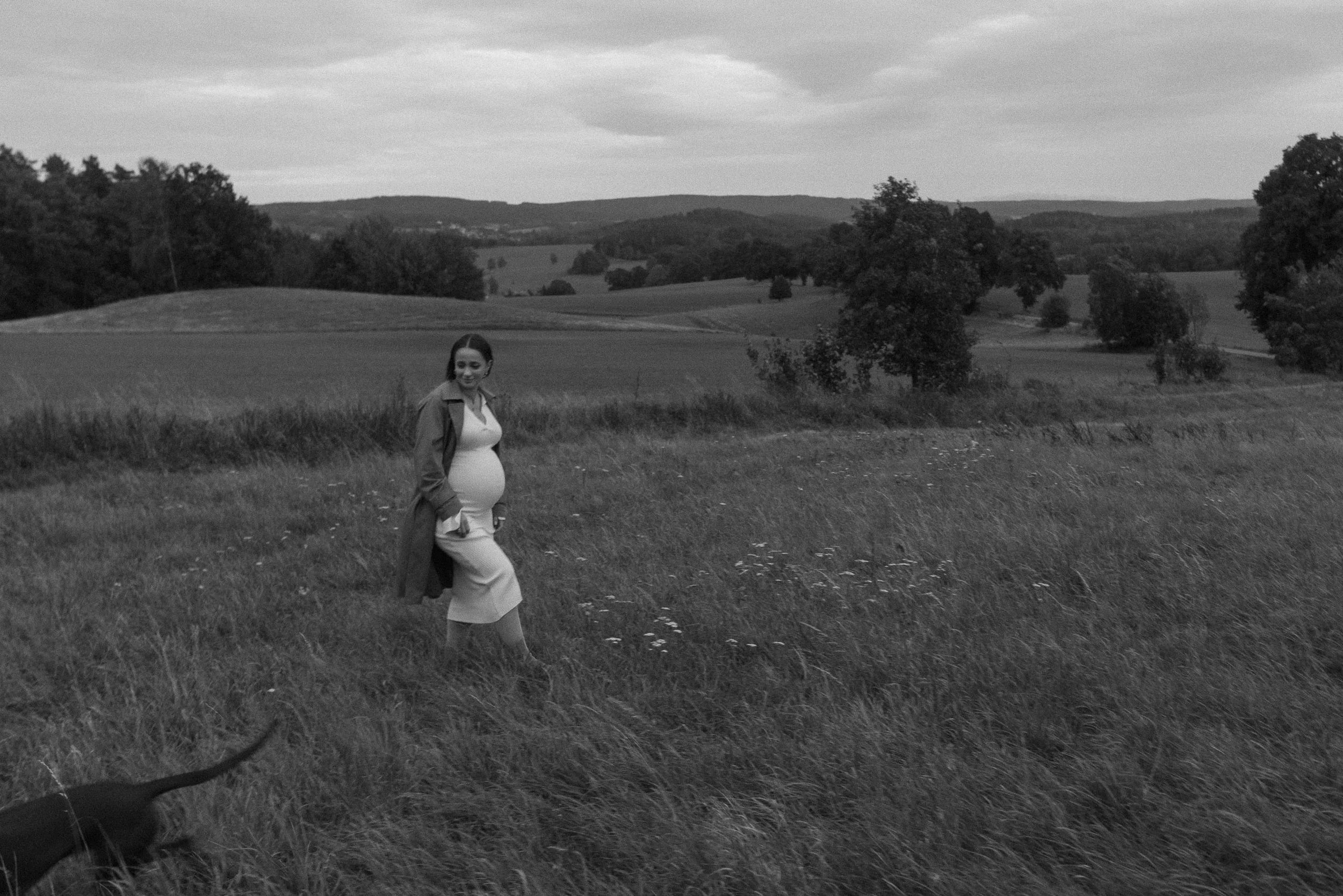 A pregnant woman walking through a grassy field with a dog, with rolling hills and scattered trees in the background, under a cloudy sky.