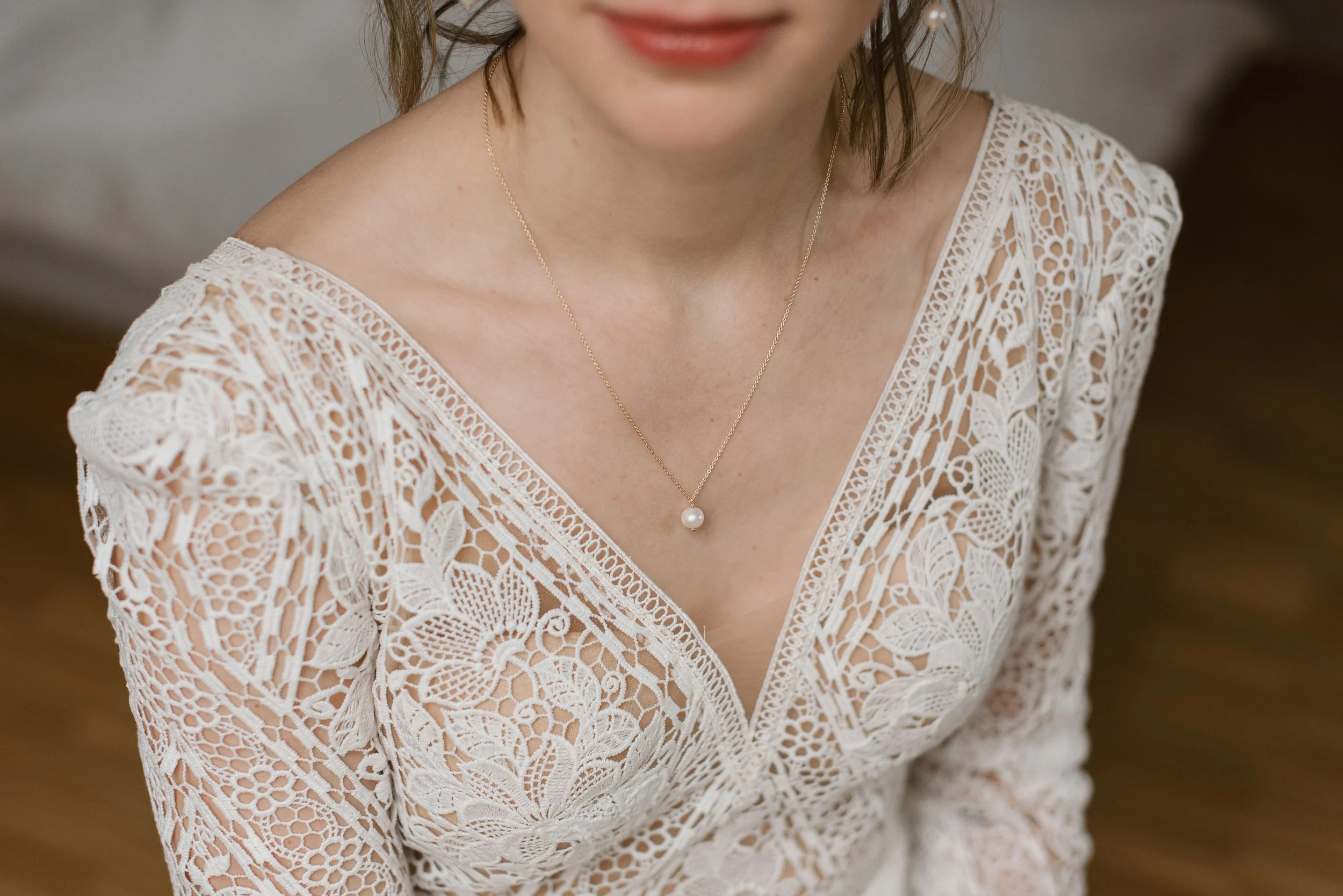 Close-up of a woman wearing a white lace dress with floral patterns, a pearl pendant necklace, and a subtle smile.