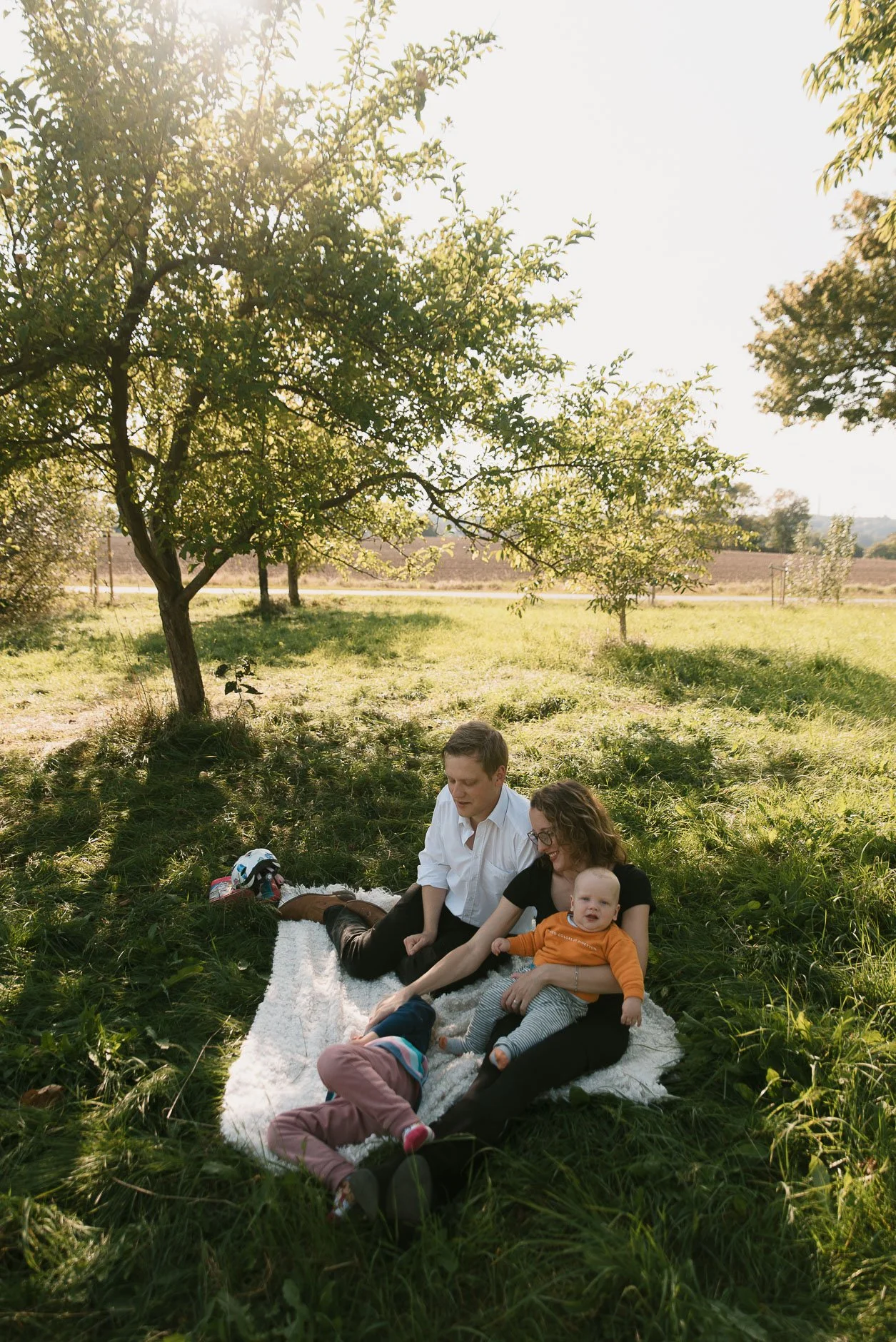 A family of four sitting on a blanket under a tree in a grassy field on a sunny day, with a man, woman, and two children, one of whom is a baby.