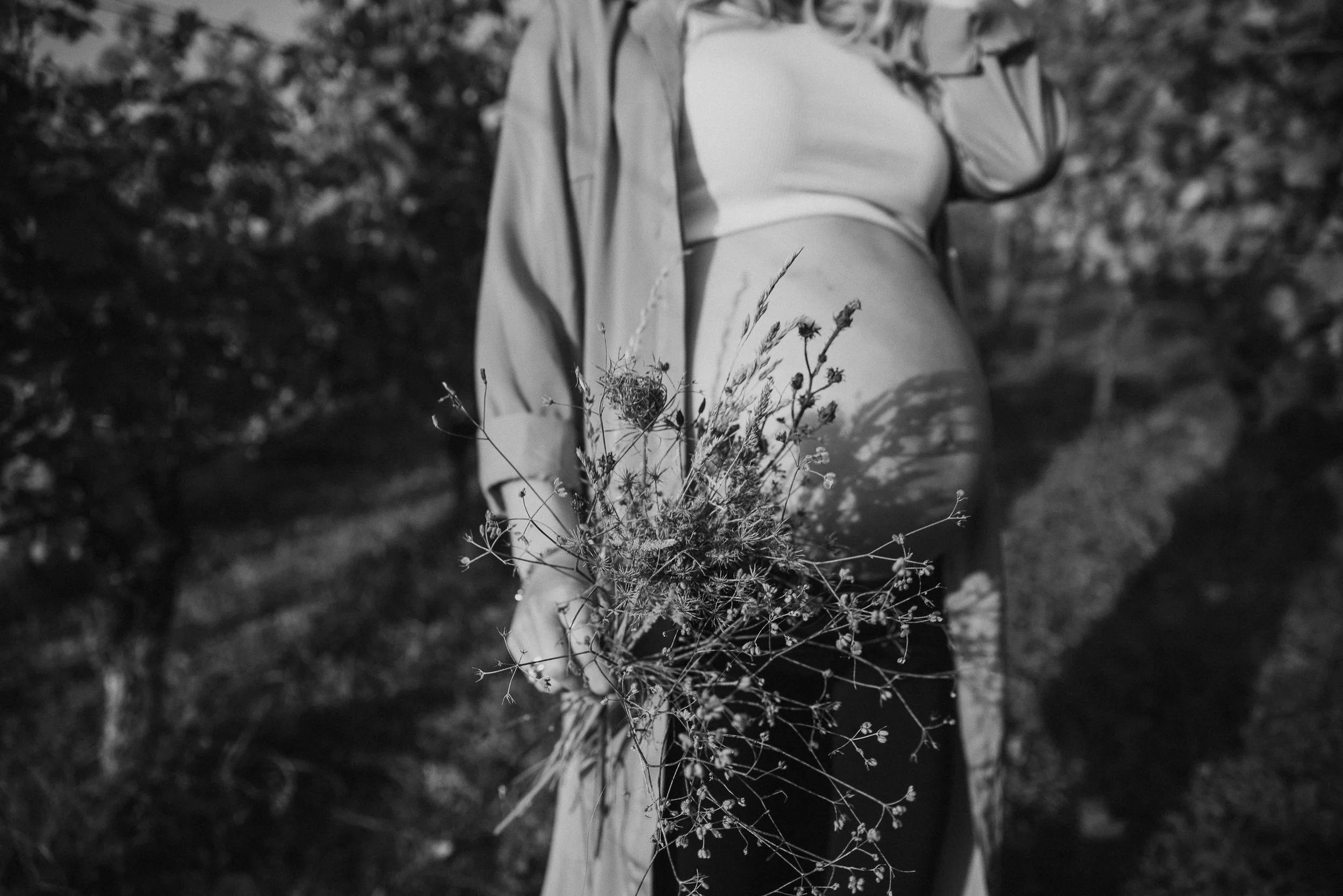 A pregnant woman holding a bouquet of wildflowers outside, dressed in a light-colored coat and a white top, with their belly visible.