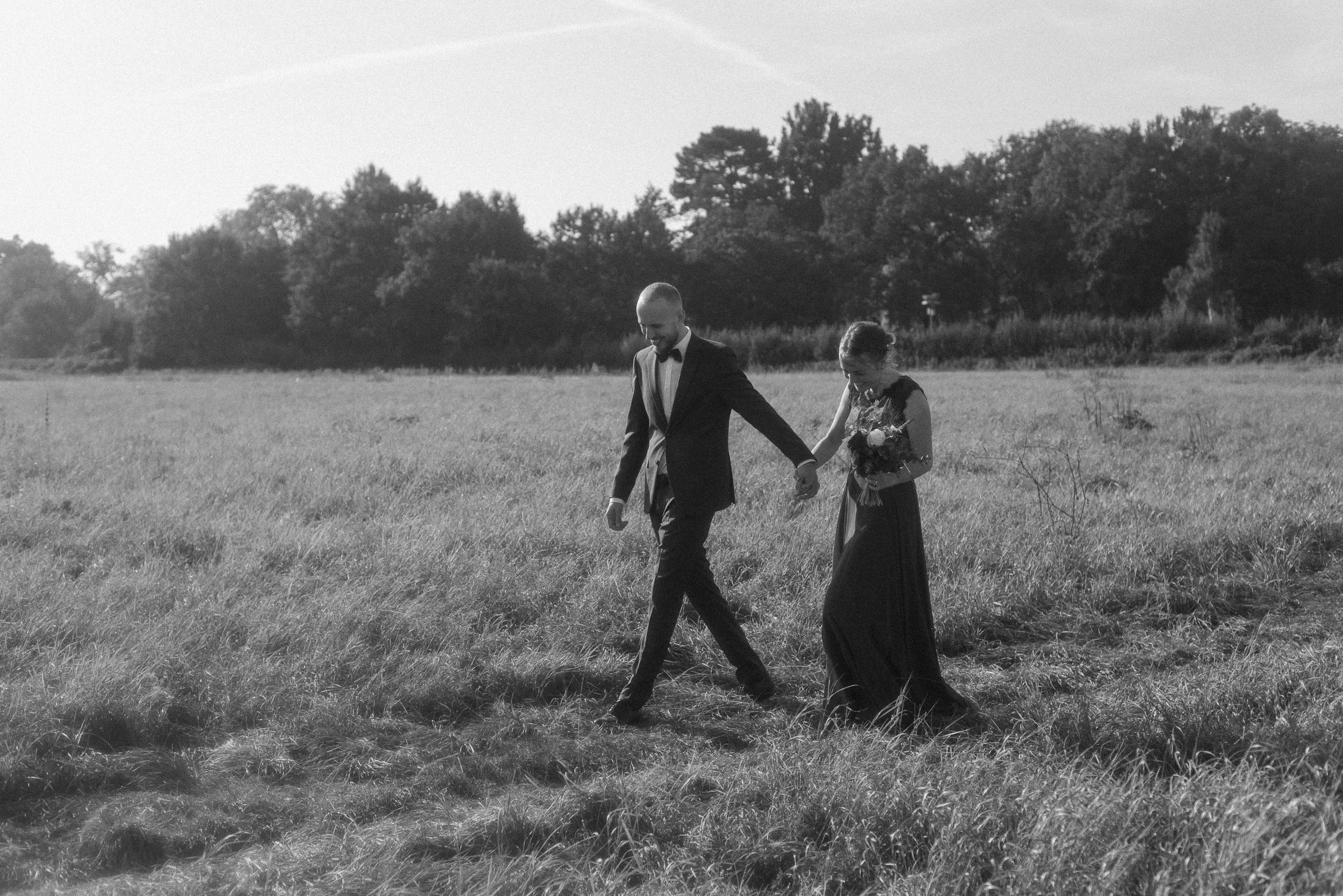 A black and white photo of a couple walking hand in hand across a grassy field during sunset, with the man in a tuxedo and woman in a formal dress holding a bouquet.