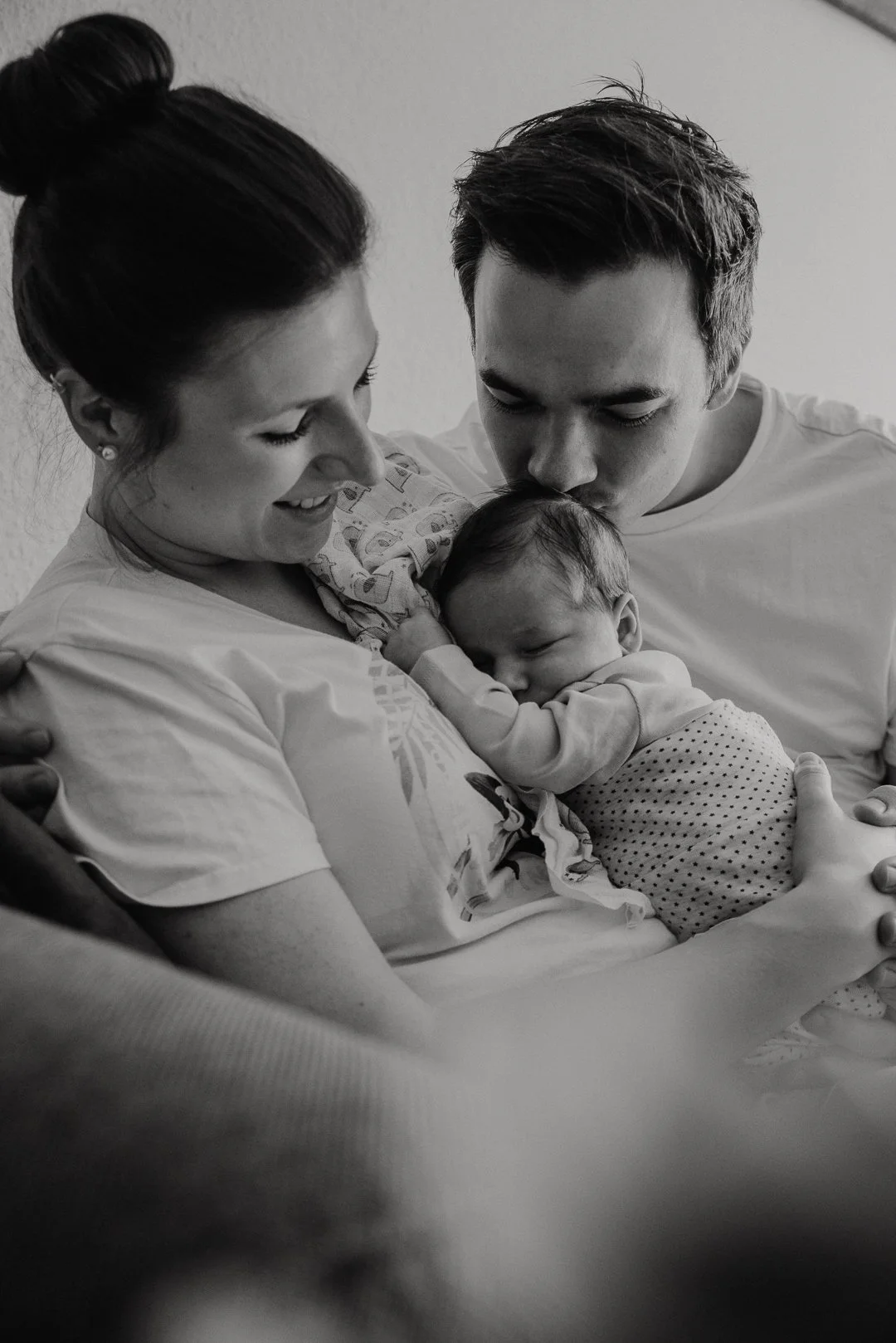 A black-and-white photograph of a family with a mother, father, and newborn baby. The mother is holding the baby close to her chest, smiling, while the father kisses the baby's forehead. The family appears to be in a hospital or home setting, experiencing a tender moment together.