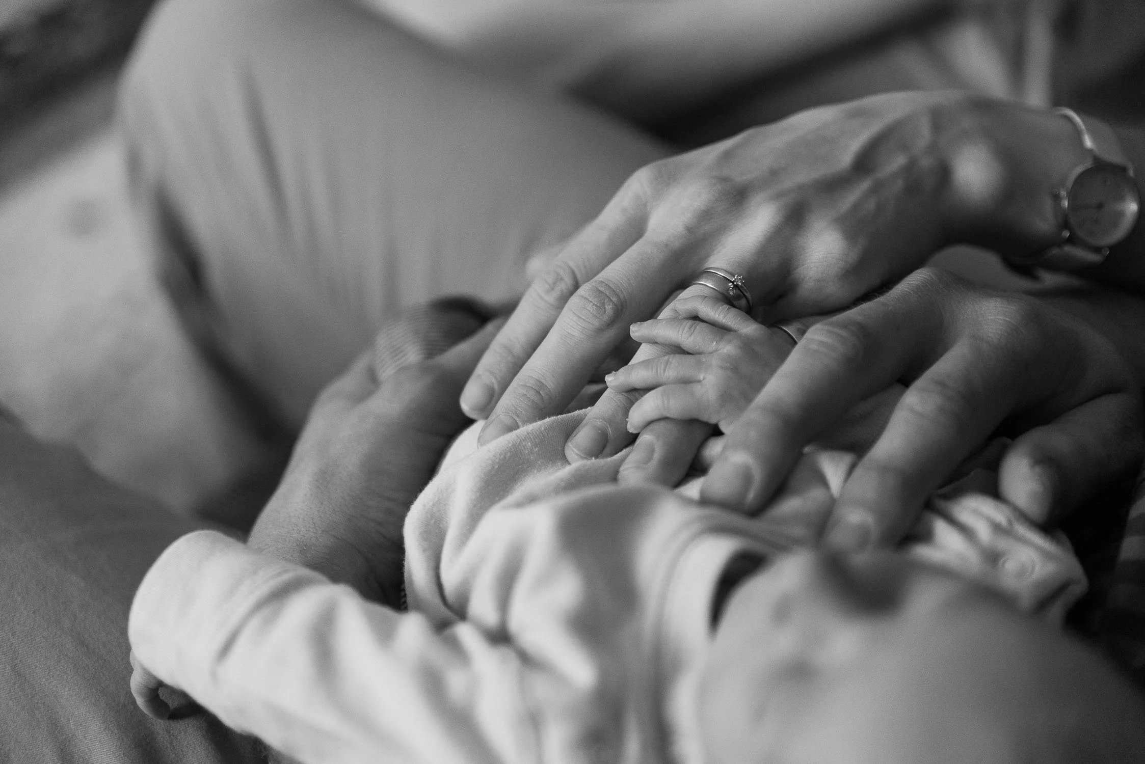 Close-up of a family holding hands, including a baby's hand, a woman's hand with a ring, and a man's hand, in black and white.