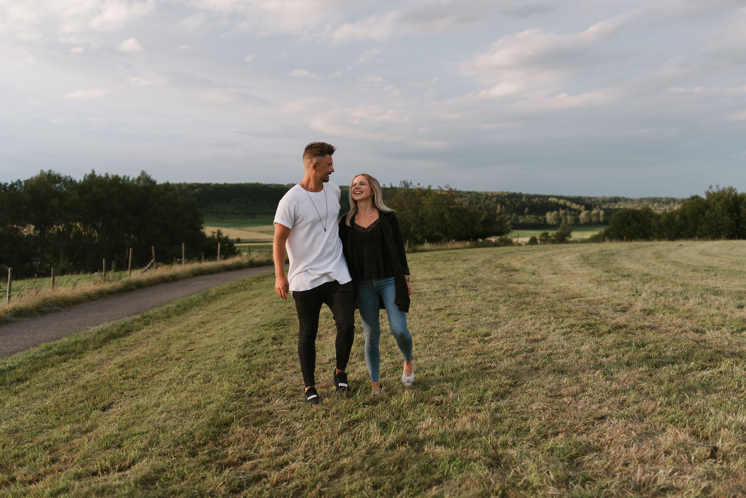 A young man and woman walking together on a grassy field in a rural area, smiling and looking at each other, with a path and trees in the background under a cloudy sky.