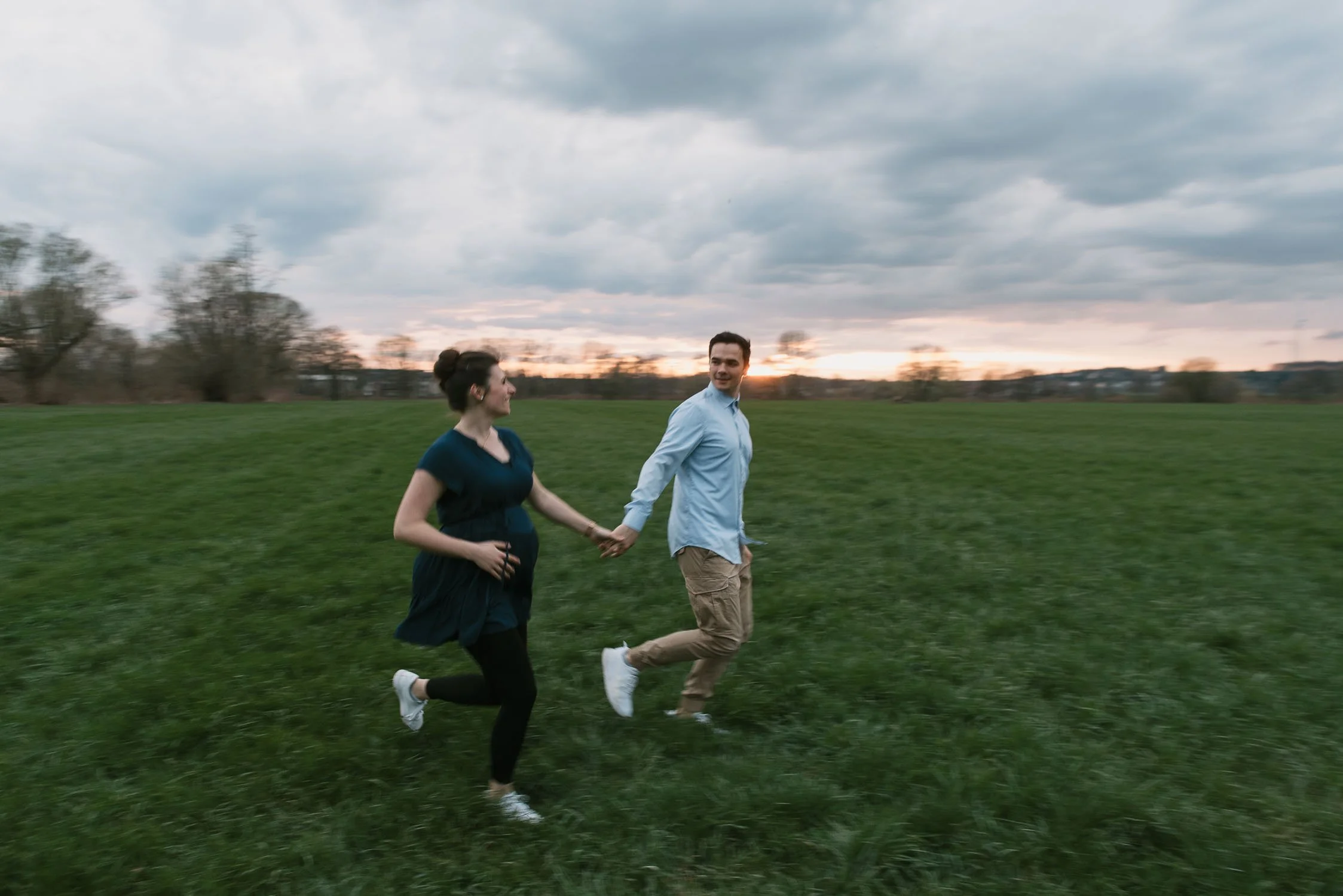 A couple holding hands and running through a green field at sunset, with a cloudy sky in the background.