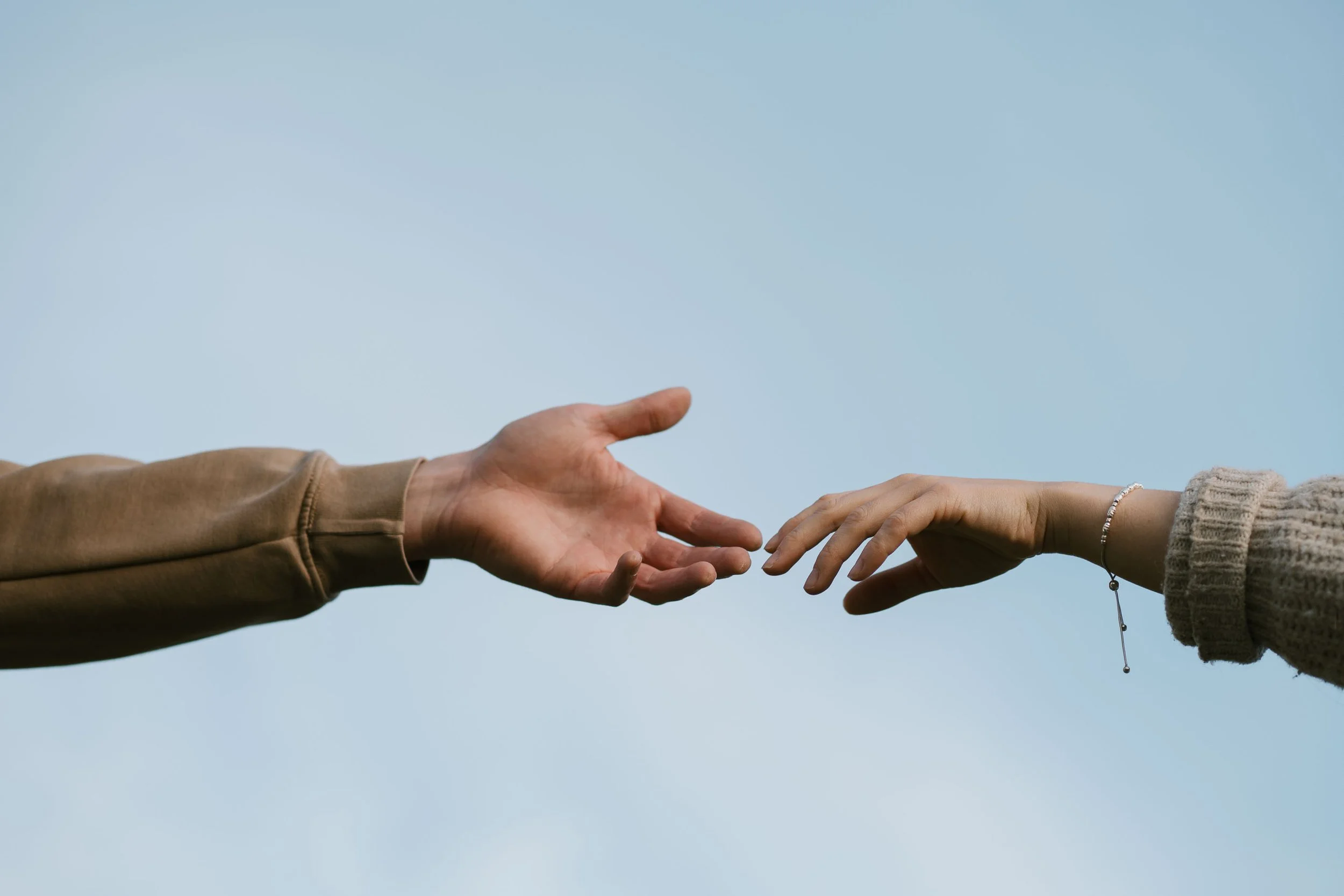 Two hands reaching towards each other against a light blue sky, one hand wearing a brown sleeve and the other a gray sweater with a bracelet.