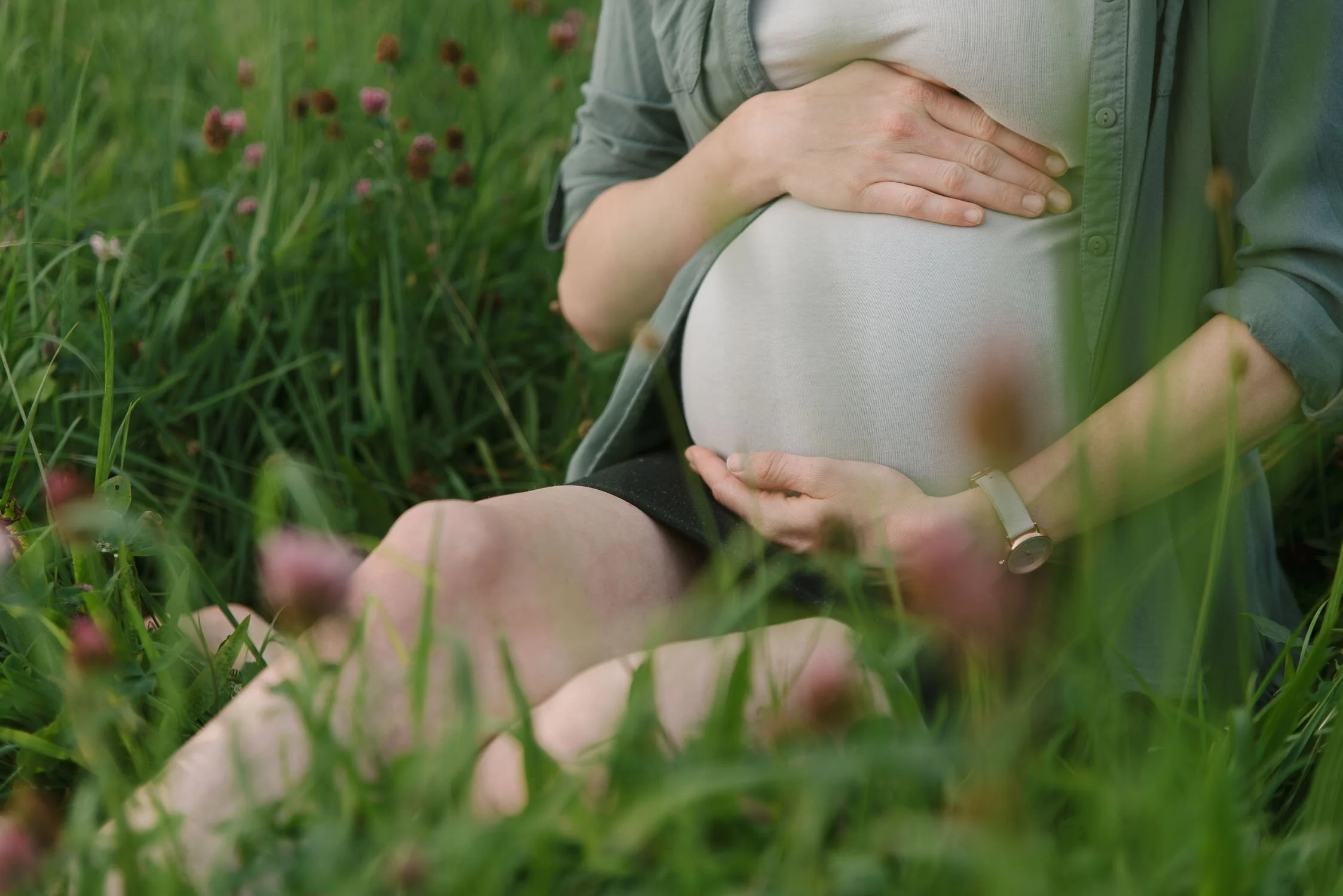Pregnant person sitting in a grassy field, holding their belly with both hands, wearing a watch and a green shirt.