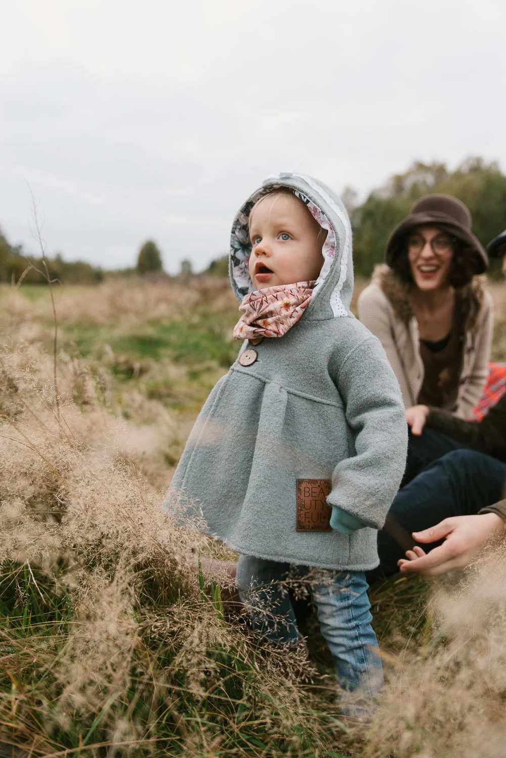 A young girl wearing a gray hooded coat and scarf stands outdoors in a natural setting with tall grass, looking curious, while a woman and another person sit in the background smiling.