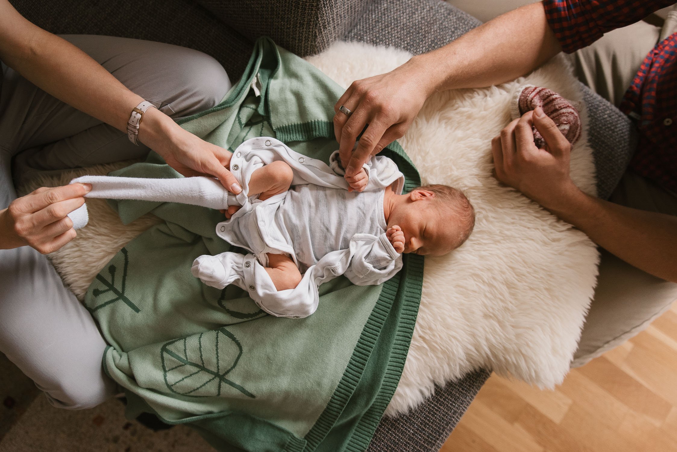 A newborn baby lying on a soft blanket with two adults gently changing the baby's sock and holding the baby's hand.