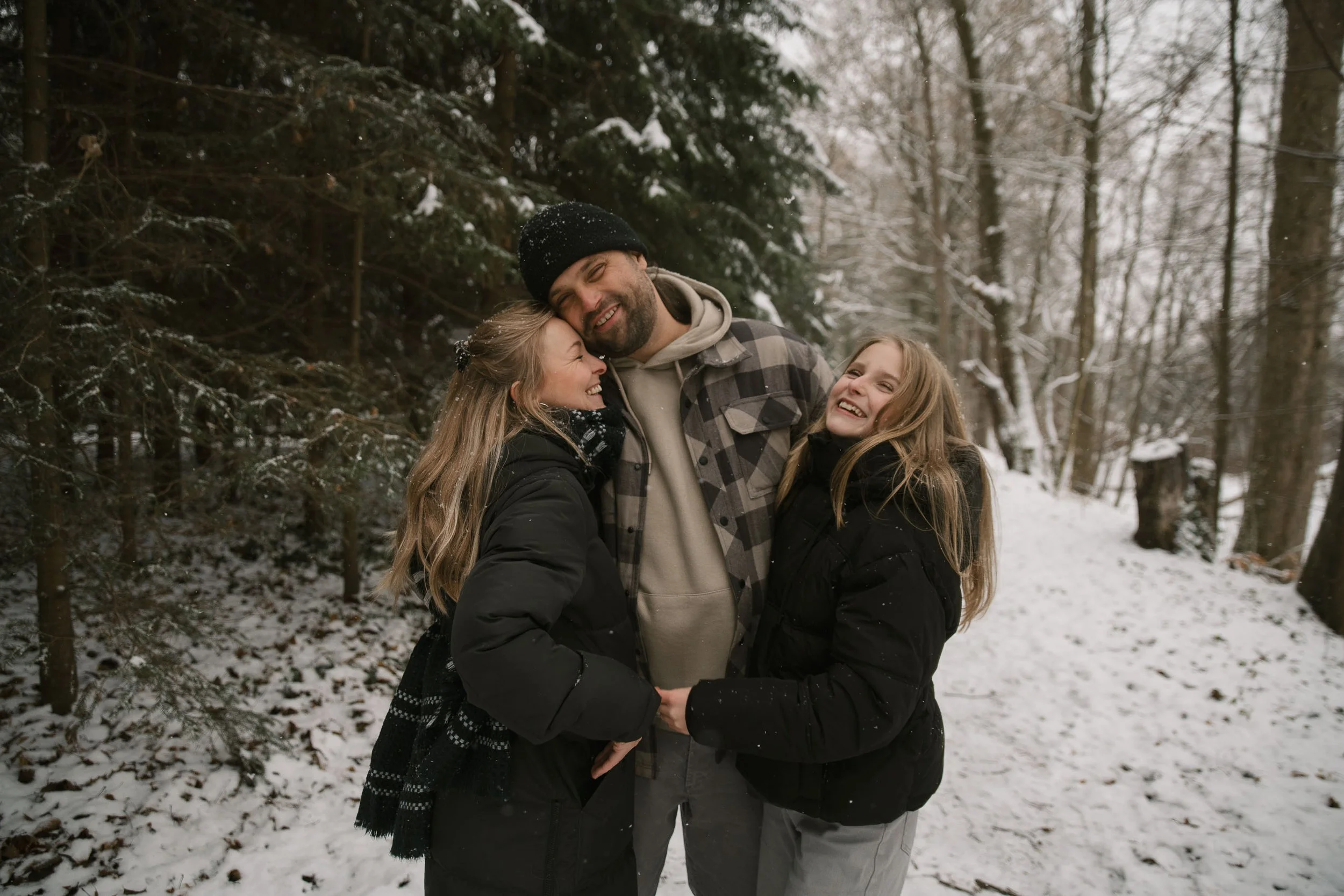 A family of three, two young women and a man, standing close together in a snowy forest, smiling and hugging each other.