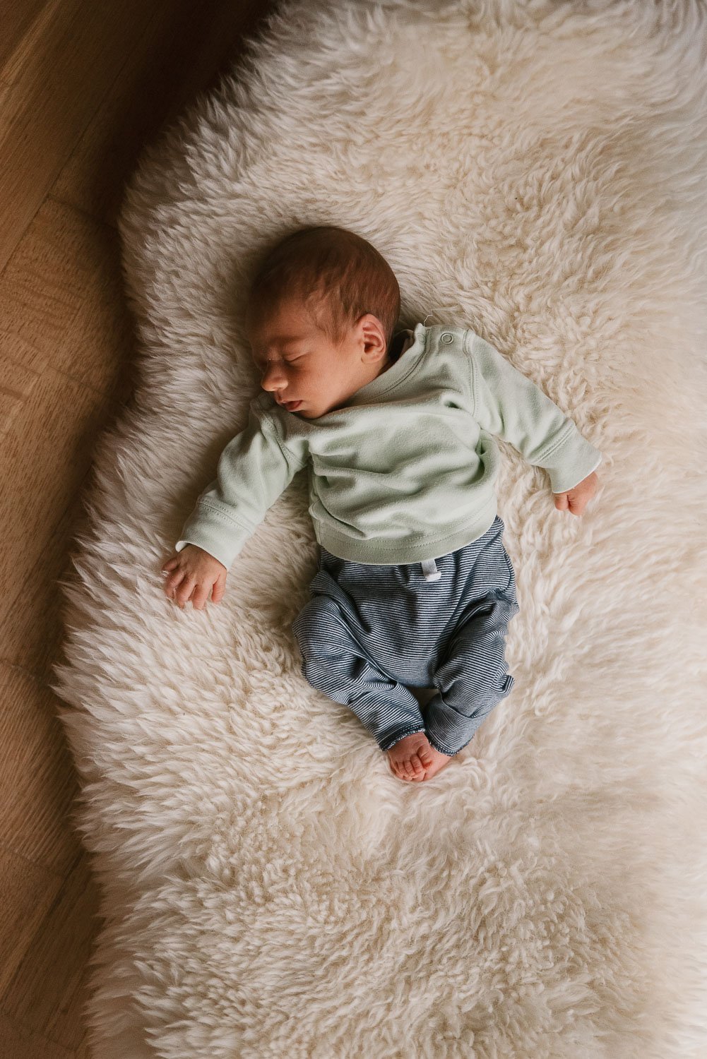A baby sleeping on a fluffy white blanket on a wooden surface, wearing a light green long-sleeve shirt and striped pants.