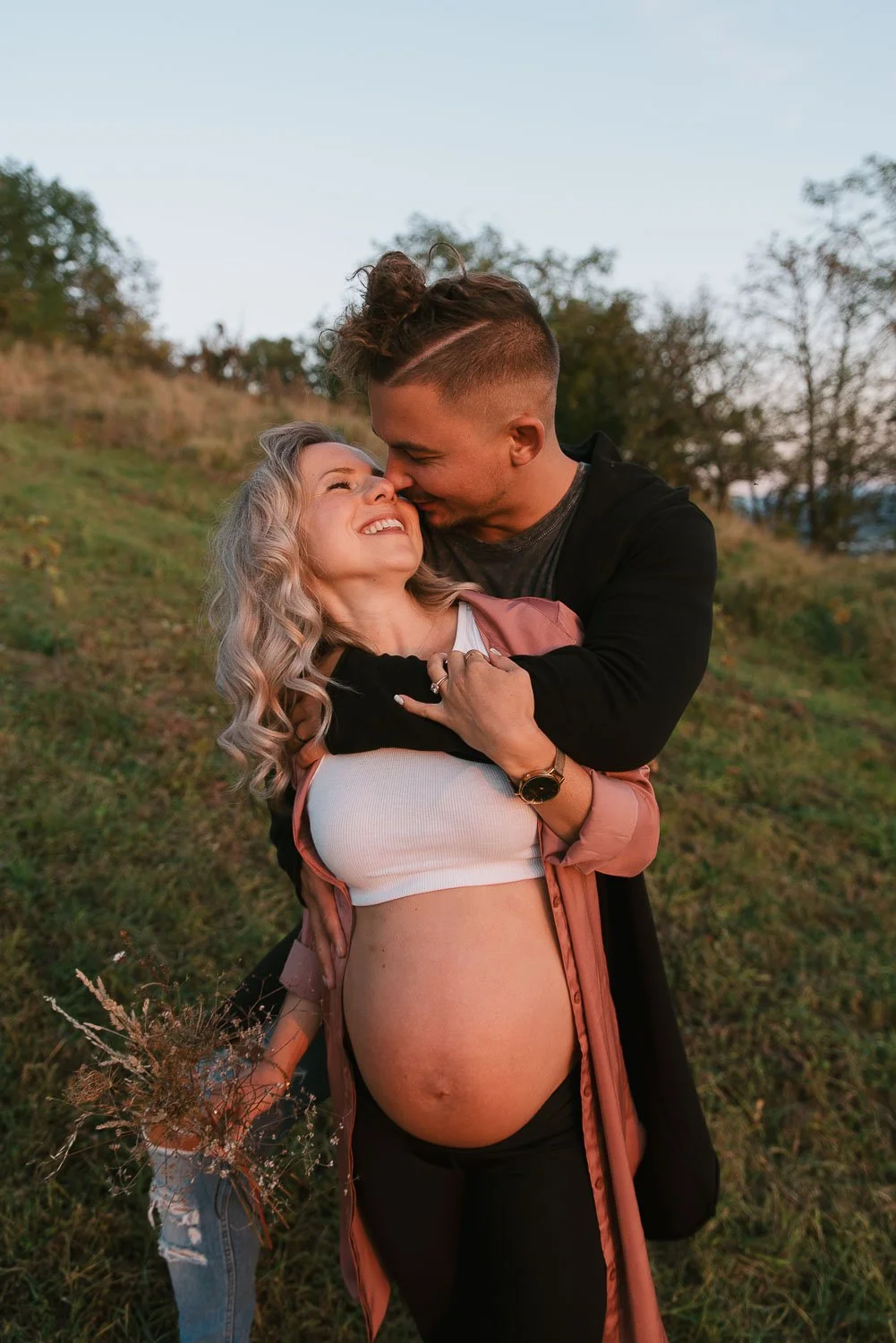 A couple, with the woman pregnant, sharing a joyful moment outdoors. The man is embracing the woman from front, touching her face, while she is smiling and looking up at him. The woman holds a small bouquet of wildflowers. The background shows greenery and trees on a clear day.
