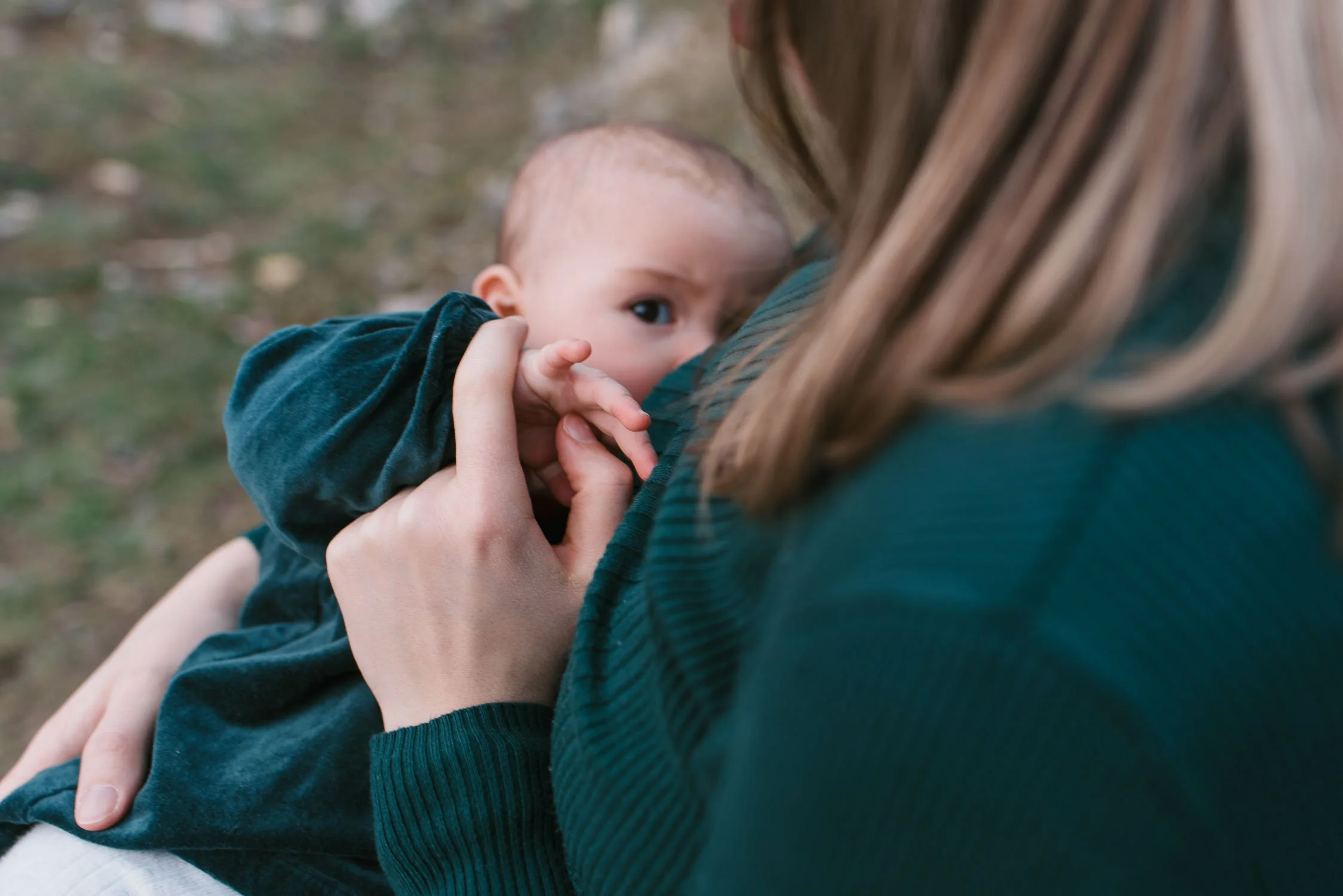 A woman holding a baby close while the baby listens and touches her face. The scene is outdoors, with blurred grass and ground in the background.