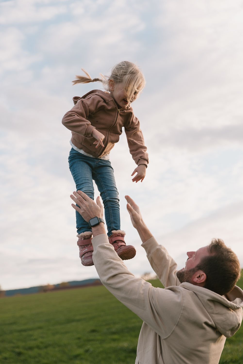 A man lifting a young girl into the air outdoors with a grassy field and cloudy sky in the background.