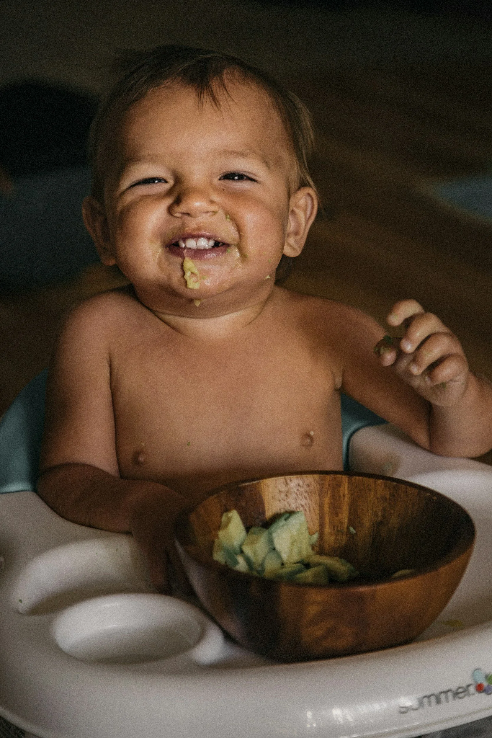 A happy young child with messy food around their mouth, holding a piece of food, sitting in a high chair with a bowl of cut fruit in front of them.