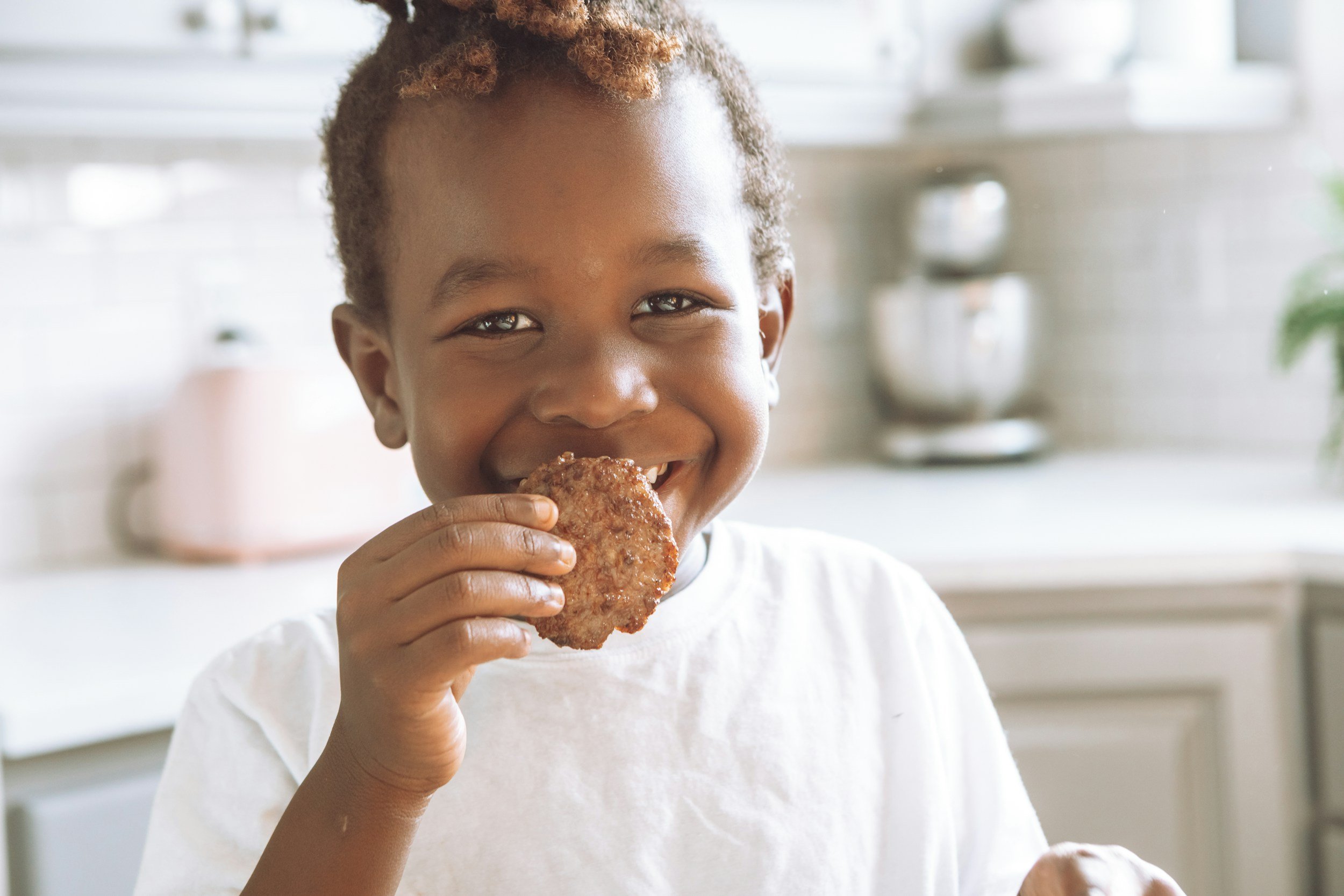 A young girl with curly hair eating a large piece of fried chicken and smiling in a bright kitchen.