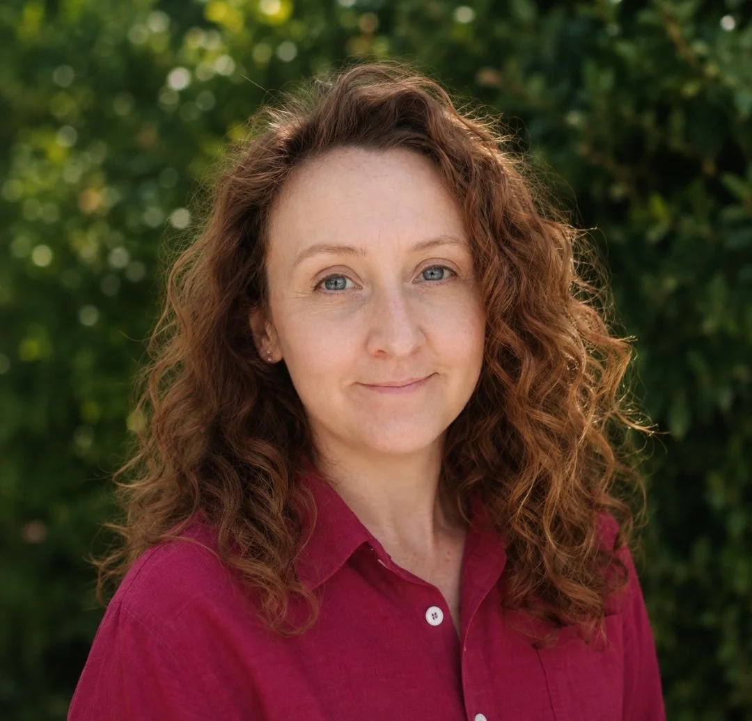 A woman with curly red hair and blue eyes, wearing a burgundy button-up shirt, standing outdoors with a background of green foliage.