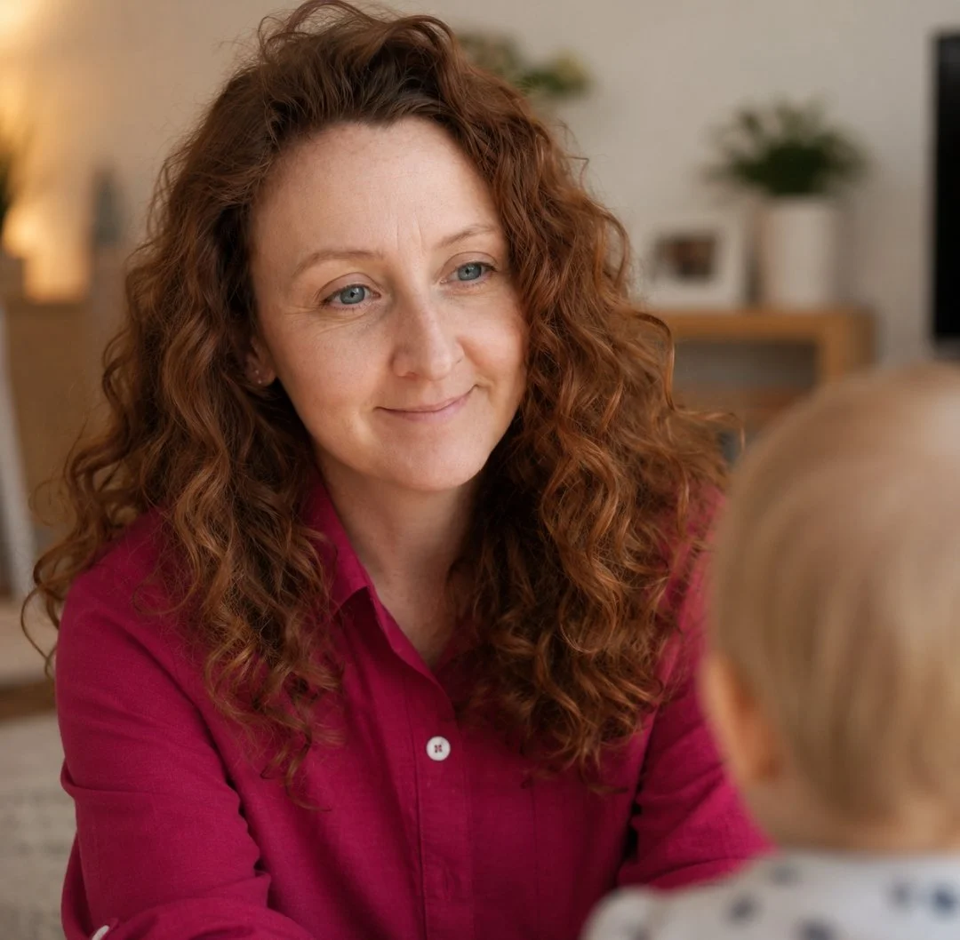 A woman with curly red hair smiling warmly at a child with blonde hair in a cozy, modern living room.