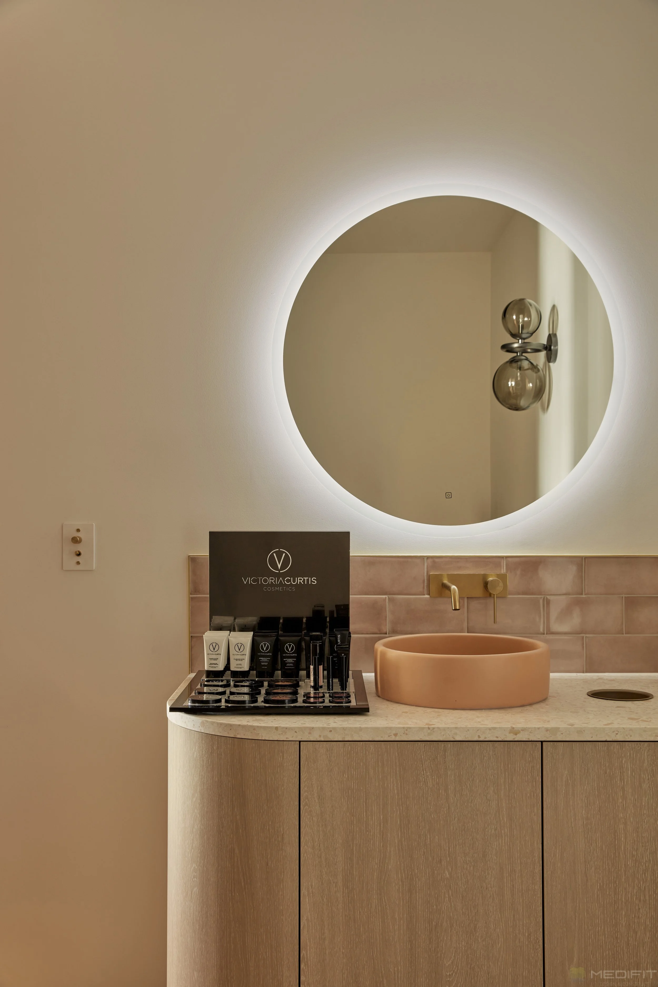 A bathroom vanity with a beige countertop, a pink circular sink, gold faucet, and a large round backlit mirror. There are skincare products on the counter, a decorative wall sculpture reflected in the mirror, and pink tiles behind the sink.