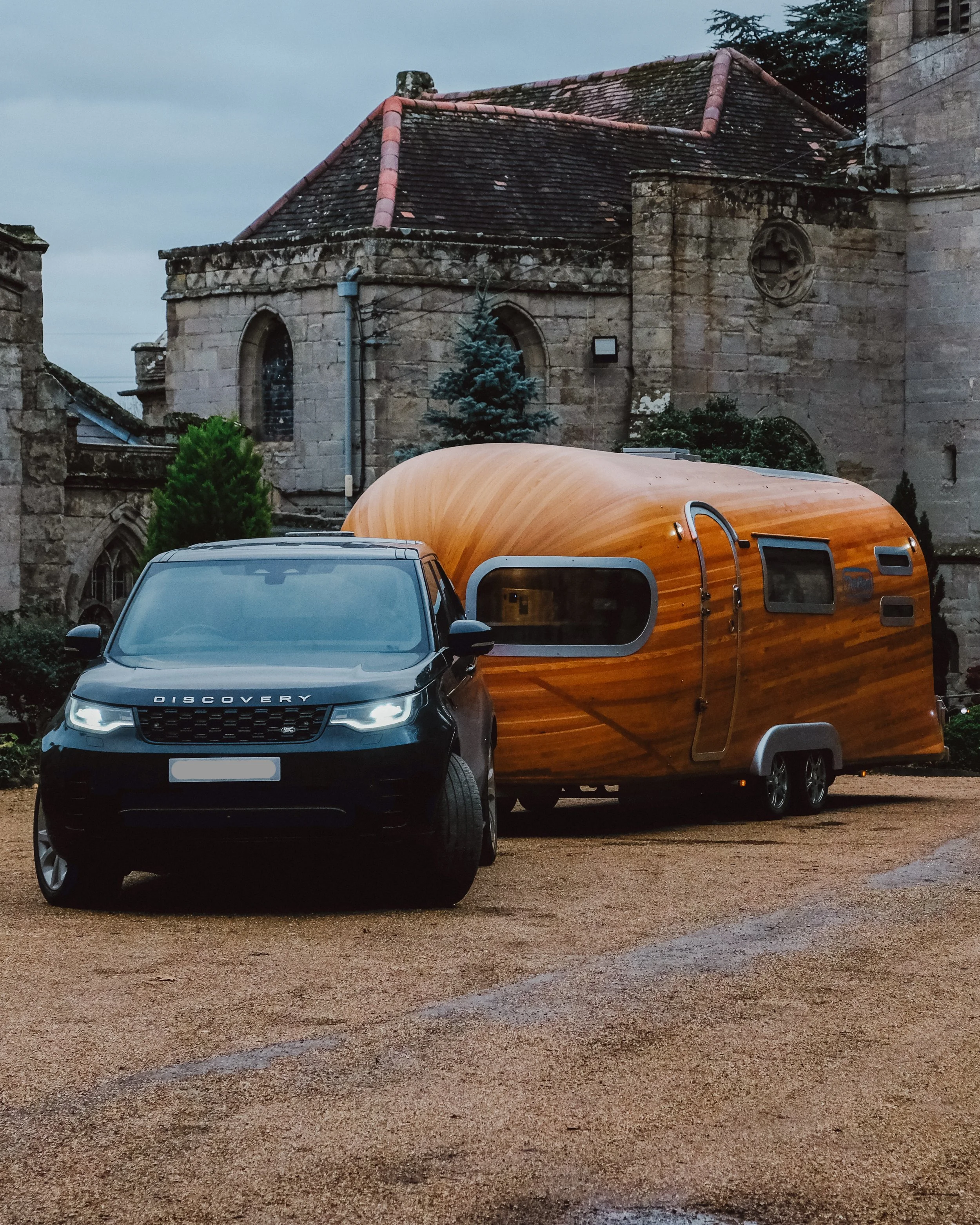A black Land Rover Discovery parked in front of an old stone church with a small caravan trailer attached. The church features arched windows, a round window, and a tiled roof, and is surrounded by trees and greenery.