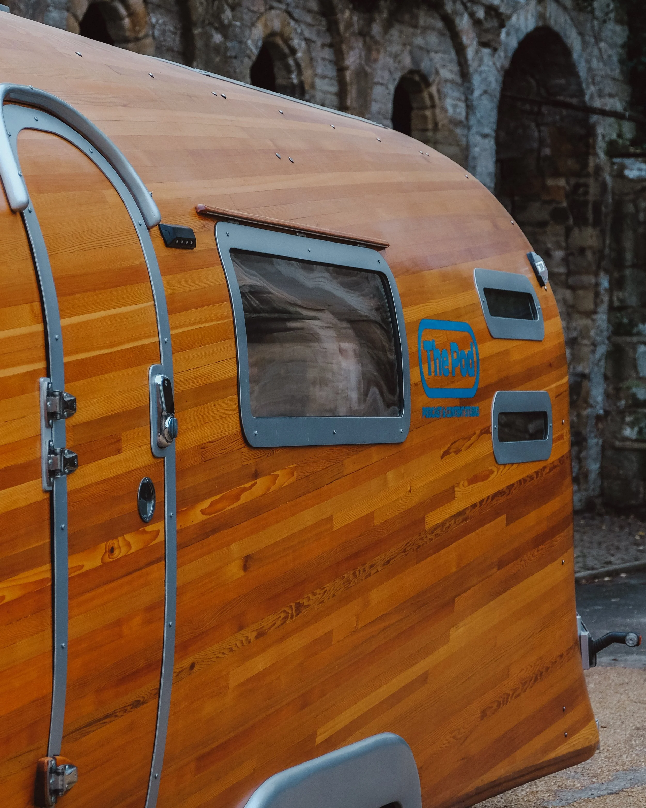 A wooden camper trailer with two windows, a door, and a logo reading "The Pod" on the side. The trailer has a curved shape and metal accents, set outdoors with a rocky background.