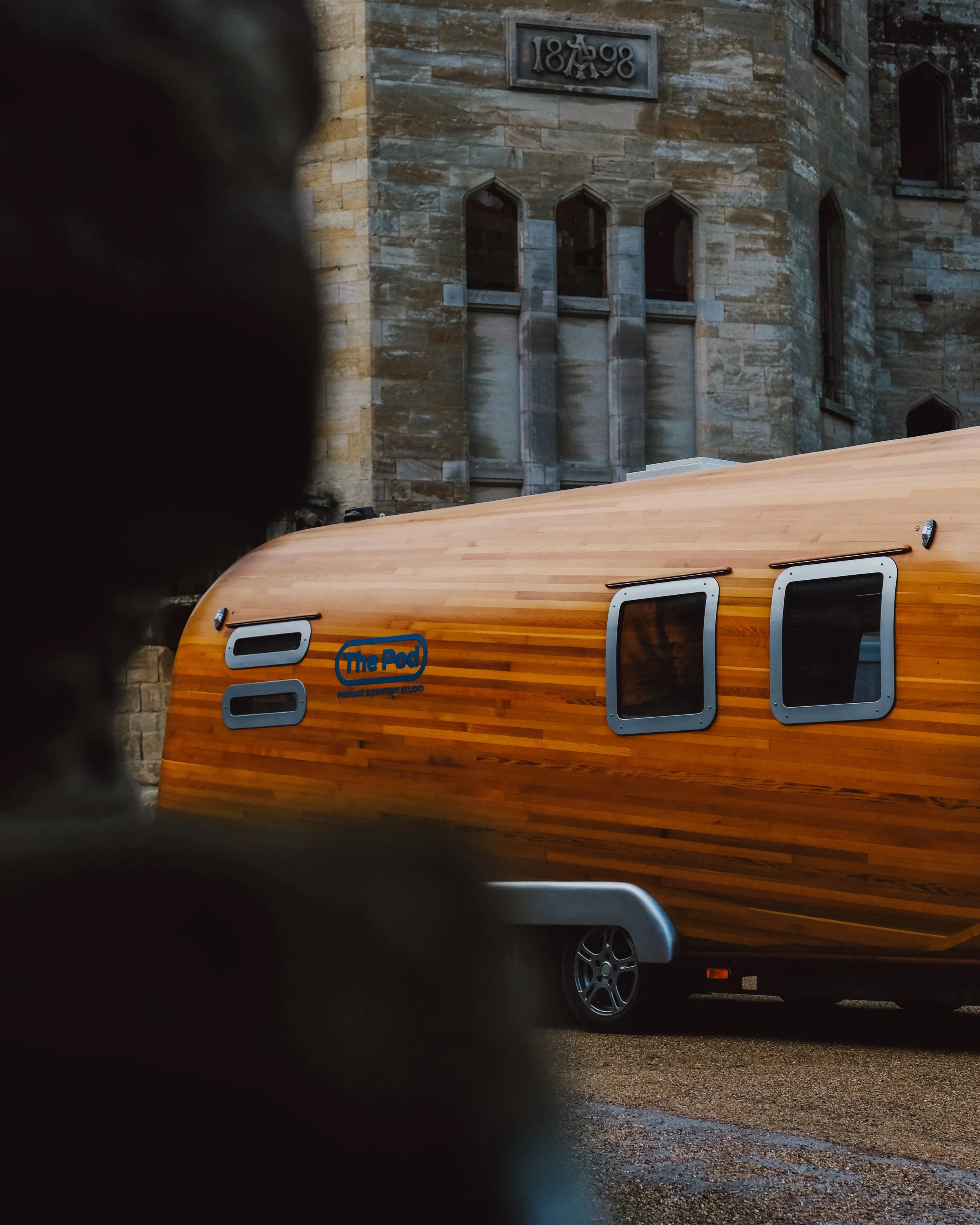 A wooden camper trailer parked on a street in front of an old stone building with a sign reading '1898'. The photo is taken from a low angle, capturing part of the street and a wheel of the trailer.