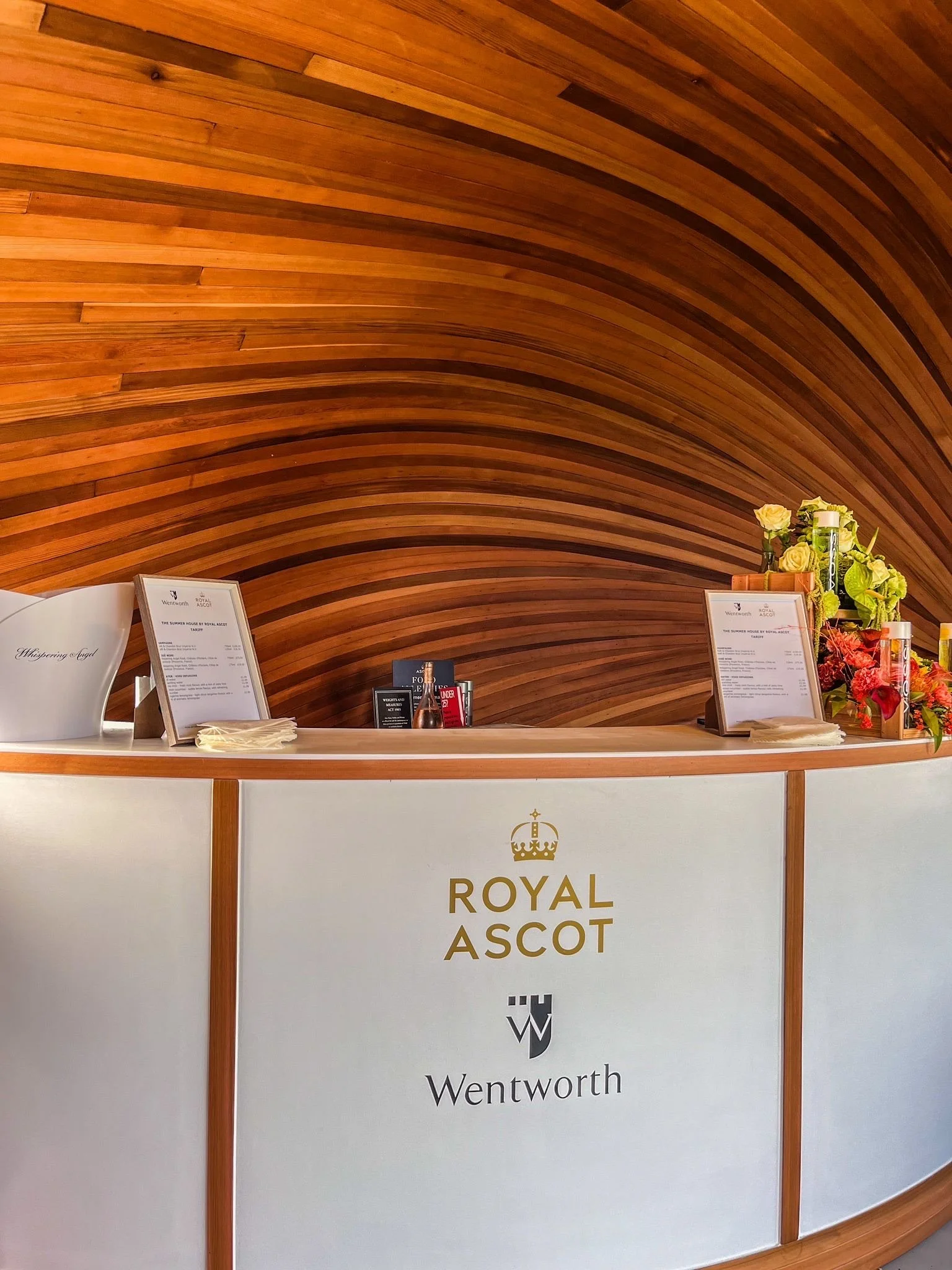 A reception desk with a sign reading 'Royal Ascot' and a crown logo, decorated with a bouquet of yellow and green flowers, against a background of curved wooden paneling.